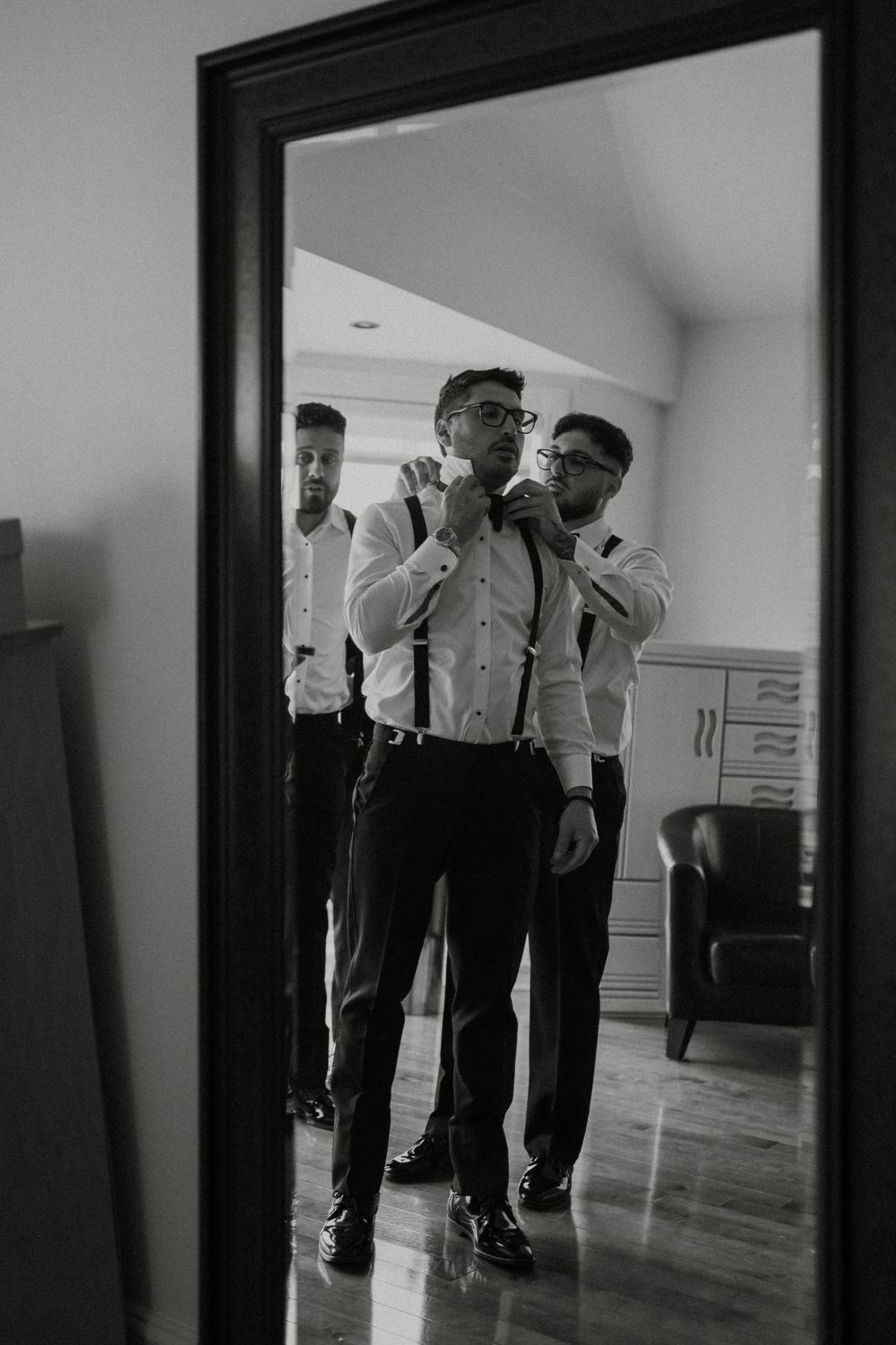 Black and white photo of groomsmen adjusting ties and suspenders in a mirror reflection.