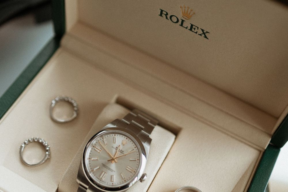 A Rolex watch sits in a cream-colored box next to two wedding rings against a light beige background.