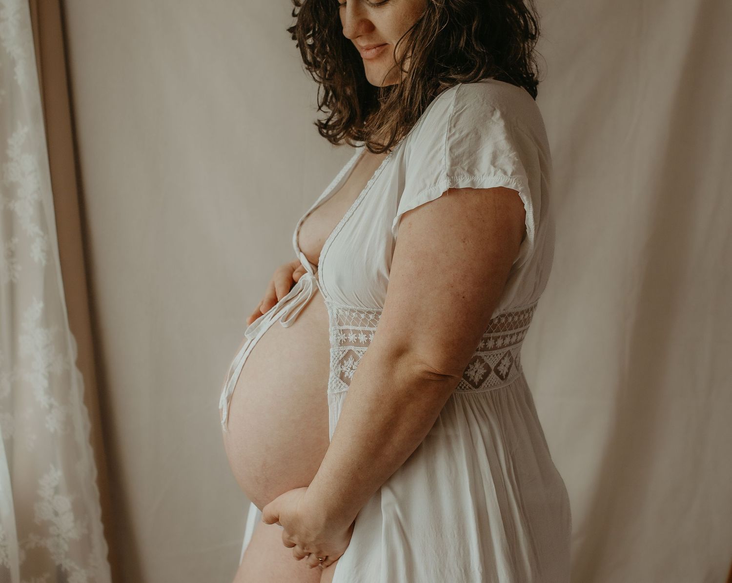Side view of an expectant mother in a white lace dress cradling her belly against a light backdrop. london and beckenham photographer.