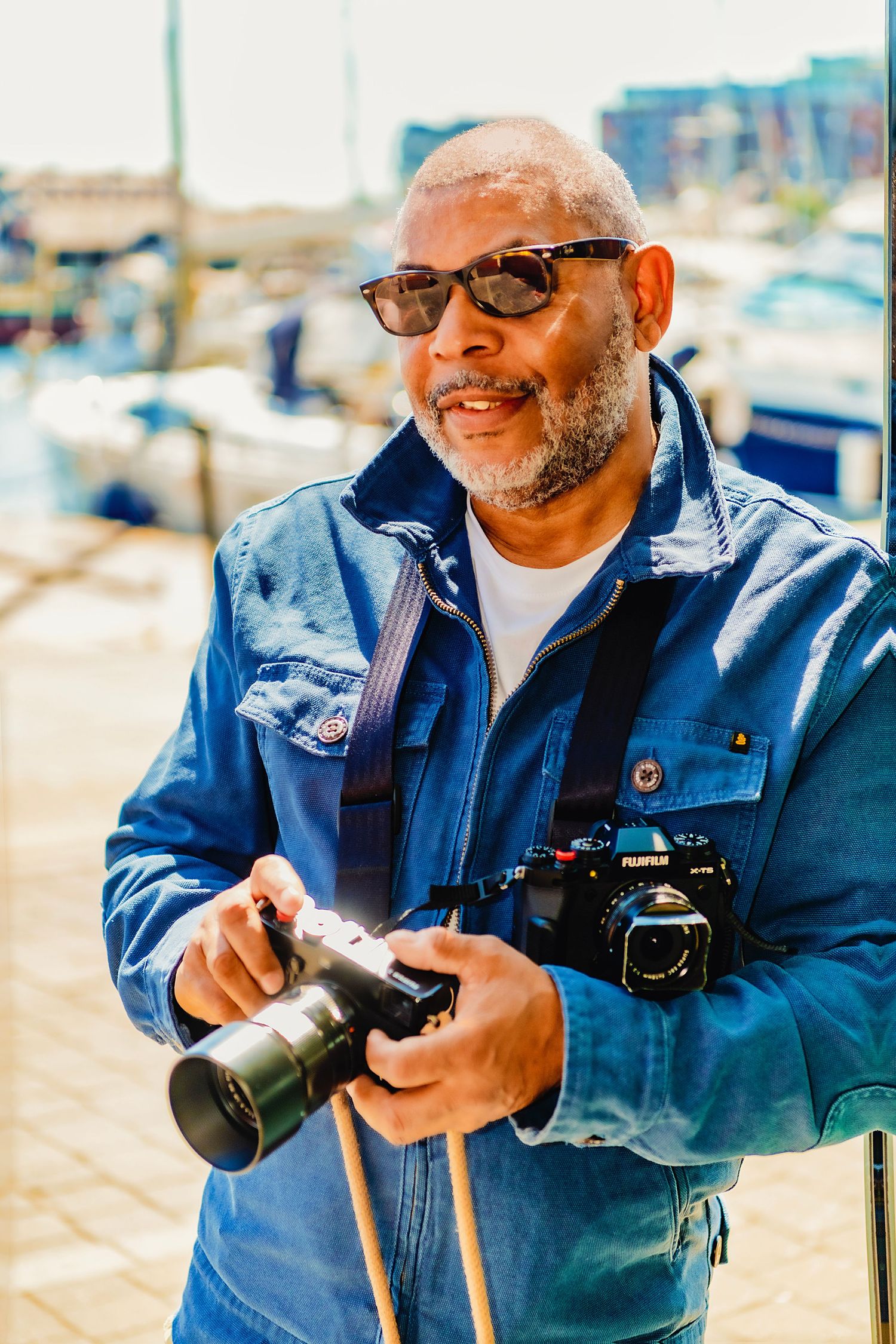 A photographer wearing a denim jacket and glasses smiles while holding camera equipment at a marina on a sunny day.
