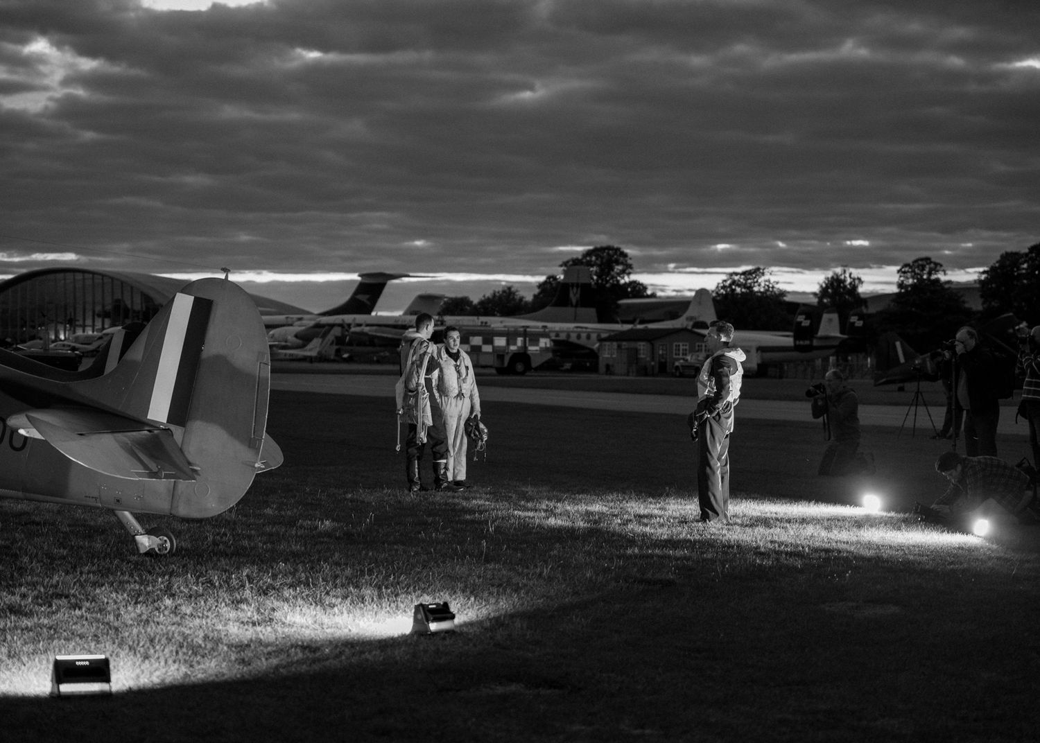 Black and white photo of vintage aircraft on airfield at dusk with ground lighting.