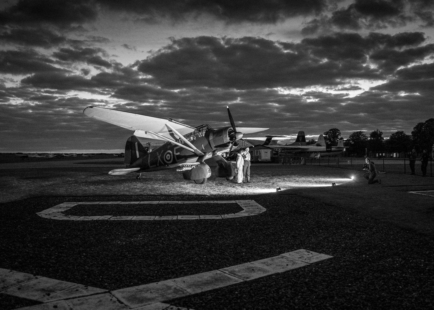 Vintage aircraft parked on airfield at sunset with dramatic cloudy sky in black and white.