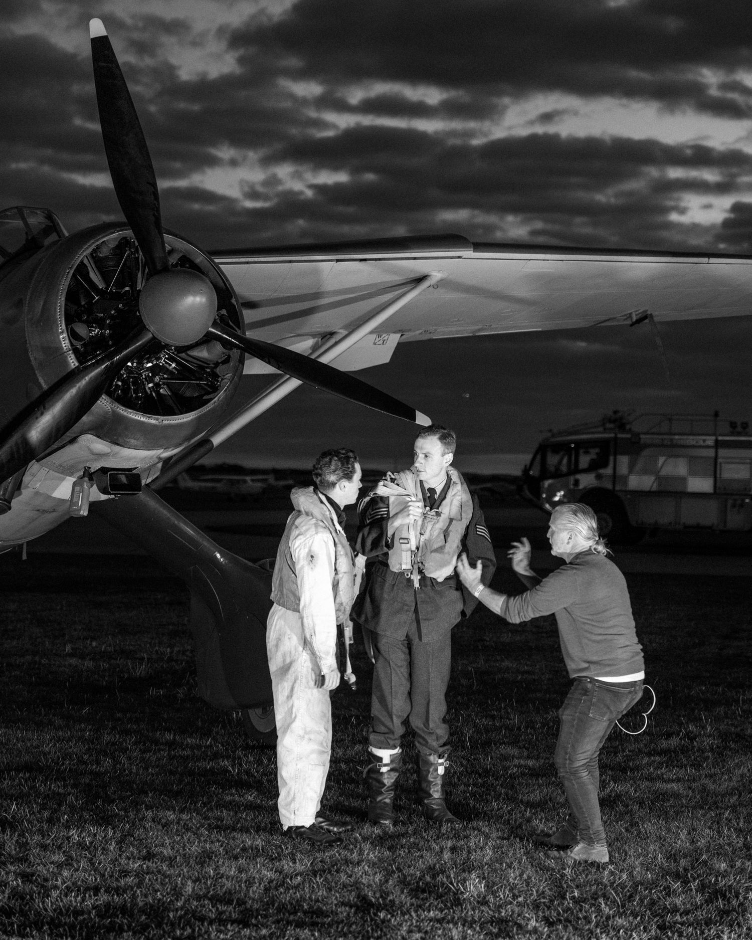 Classic propeller aircraft on airfield with ground crew gathered around engine at dusk.