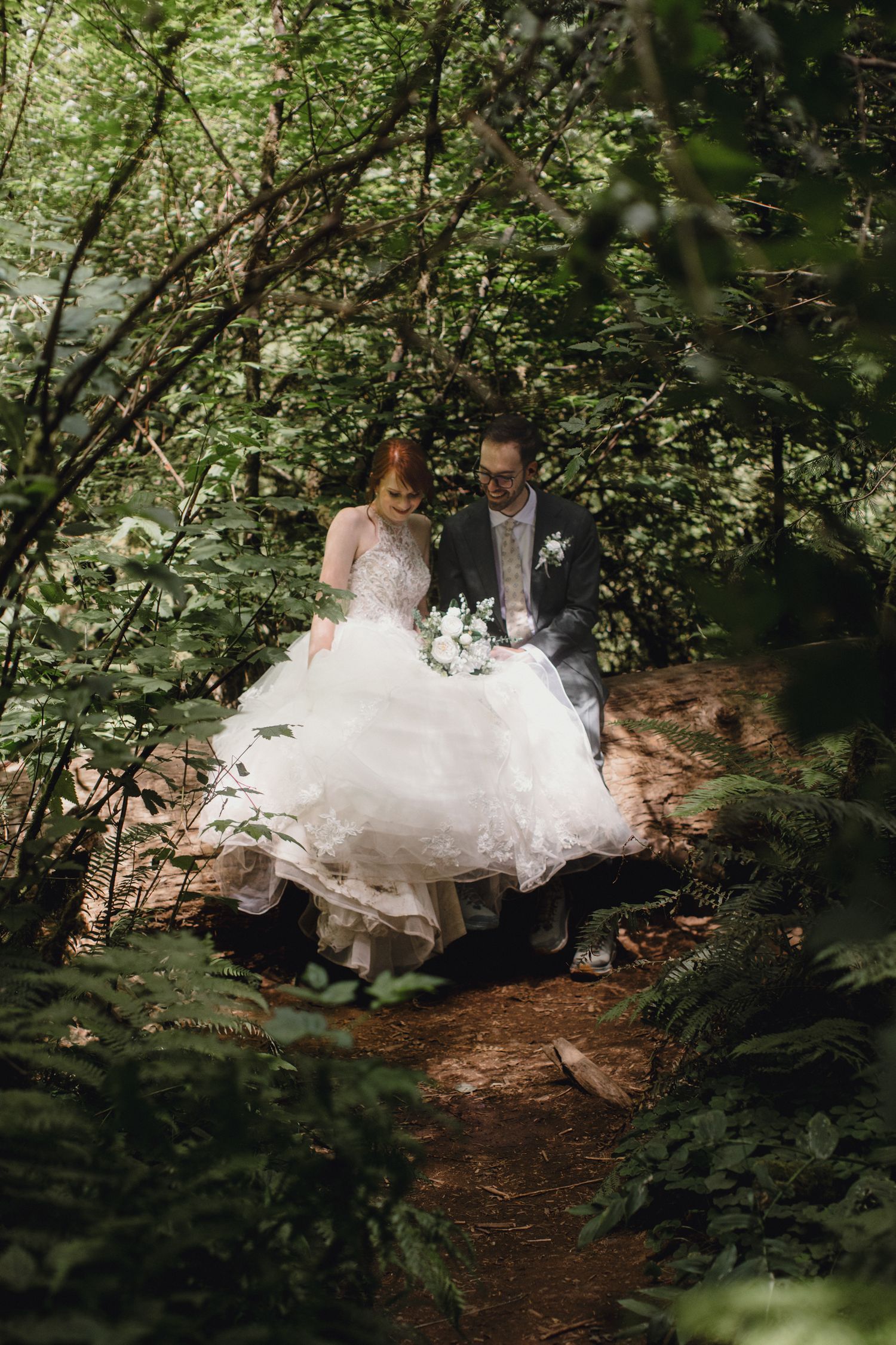 A couple in wedding attire sits on a log in a lush forest setting surrounded by green foliage.