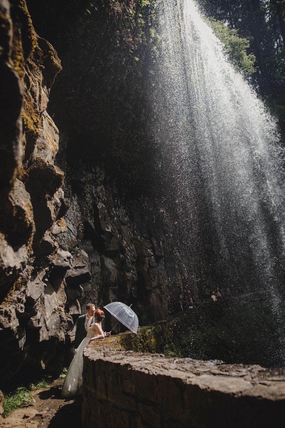 Person holding umbrella stands beneath dramatic waterfall cascading down rocky cliff in moody natural scene.