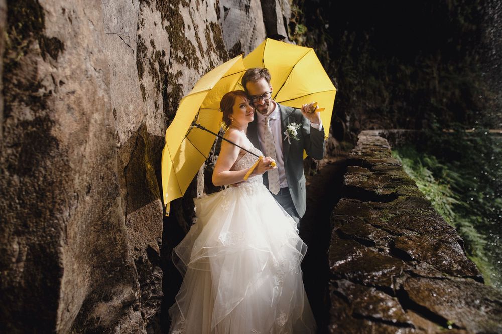 Couple in wedding attire stands beneath yellow umbrella next to dramatic rocky cliff wall.