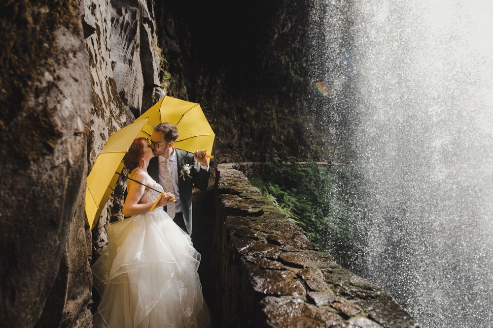 A couple with a bright yellow umbrella stand behind a cascading waterfall on a rocky ledge while taking engagement photos.