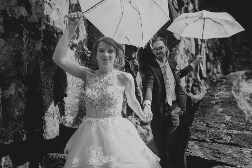 Bridal party holding white umbrellas and walking down a garden path during an outdoor wedding celebration.