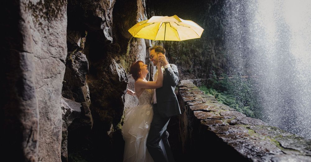 A couple with an umbrella navigates along a stone walkway in the rain near a cascading waterfall.
