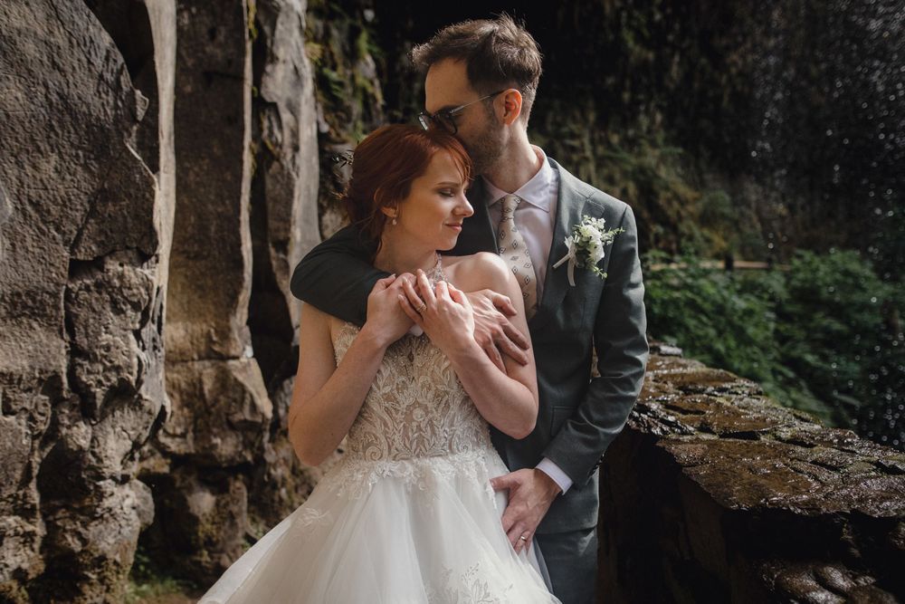 A romantic wedding couple embraces against a stone wall backdrop surrounded by natural greenery.