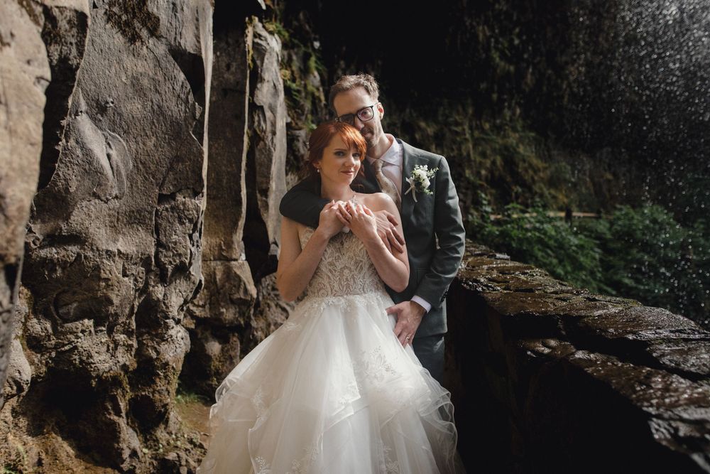 A couple in wedding attire share an intimate moment near a rocky cliff wall in a natural outdoor setting.