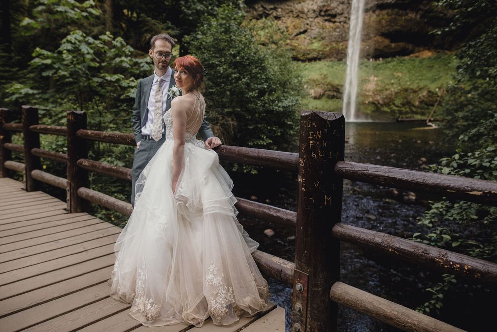 Newlyweds stand together on a rustic wooden bridge surrounded by lush greenery and a waterfall.