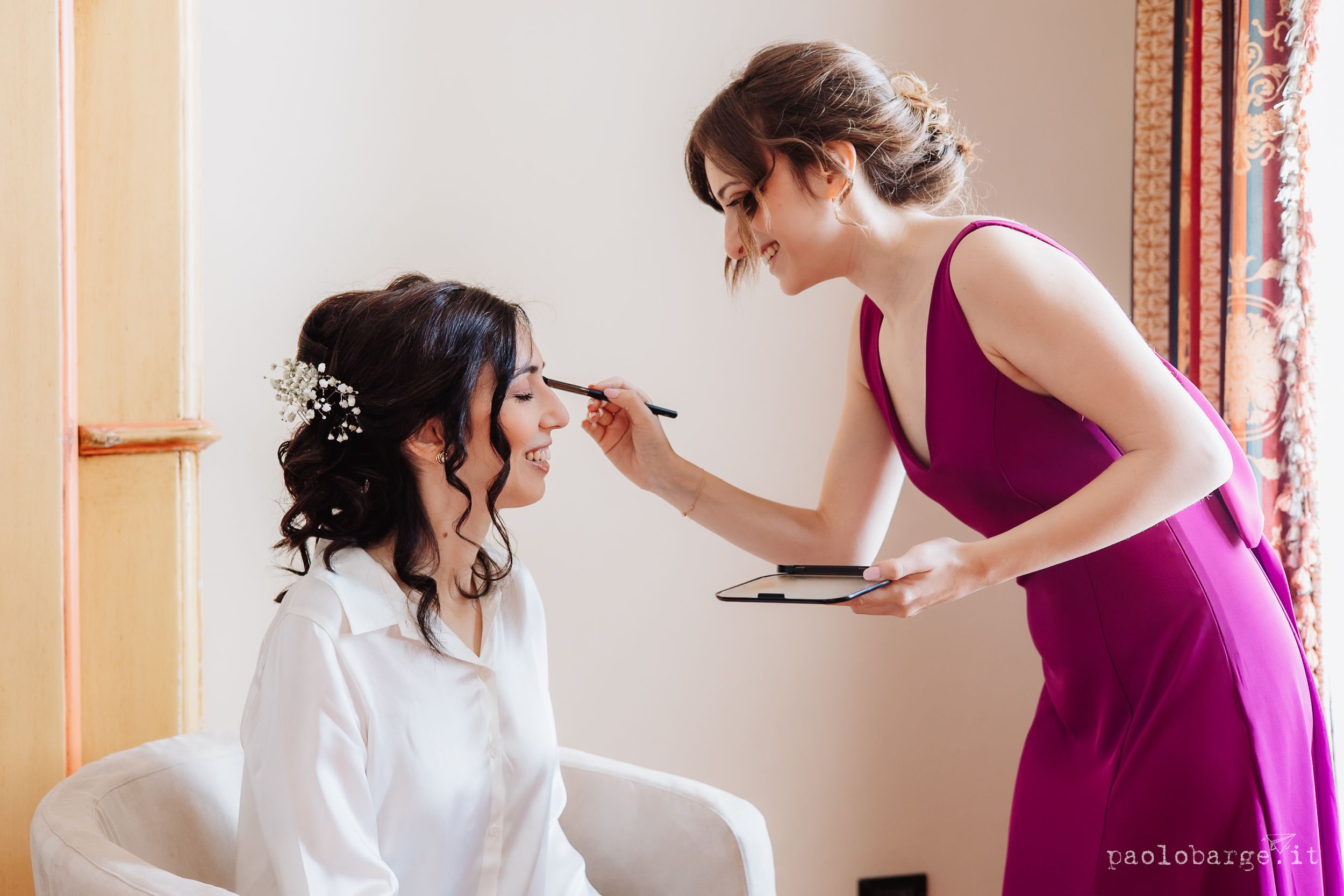 A makeup artist in a burgundy dress applies cosmetics to someone wearing a white robe during wedding preparations.