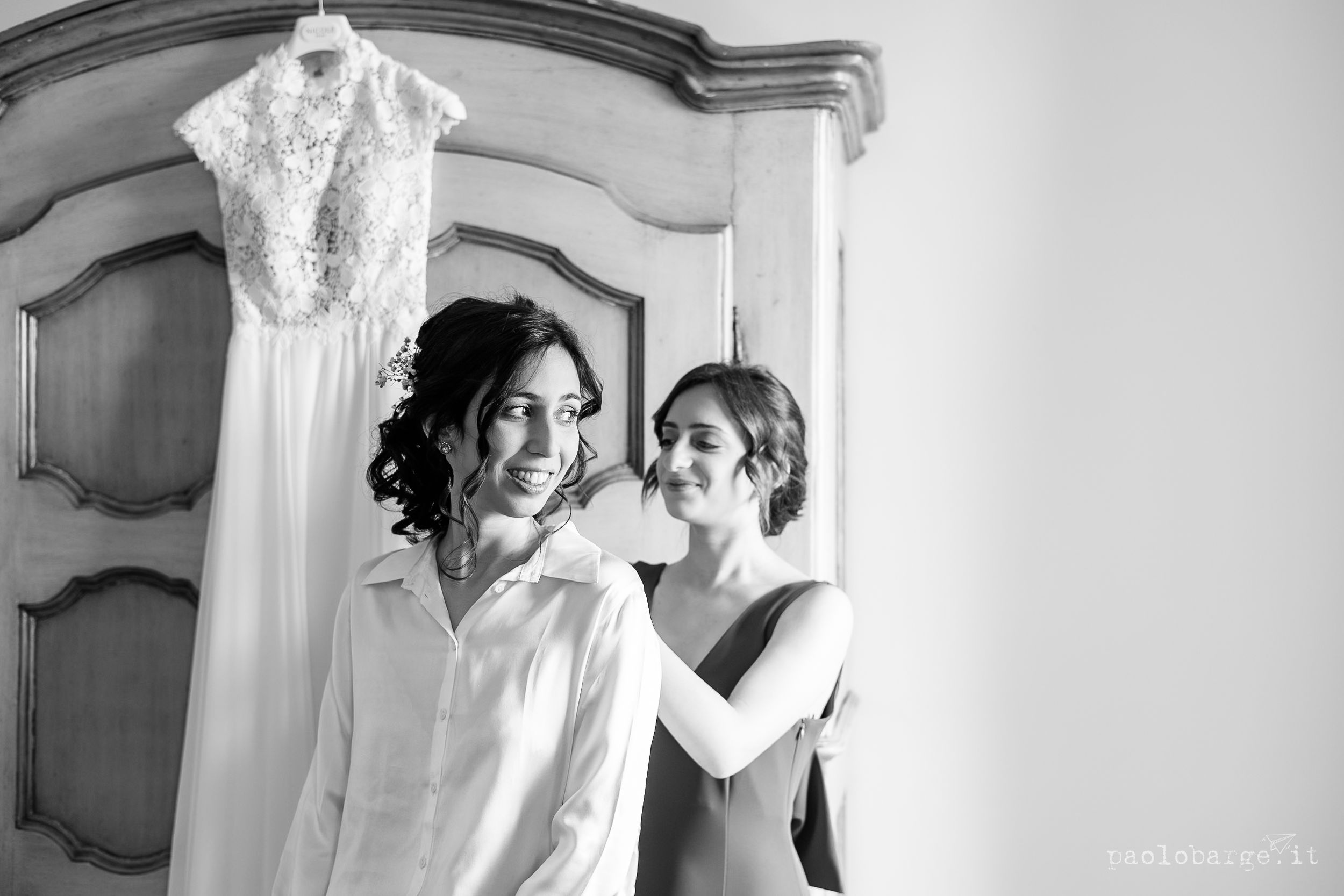 Black and white photograph showing wedding preparations with a white lace dress hanging in the foreground.
