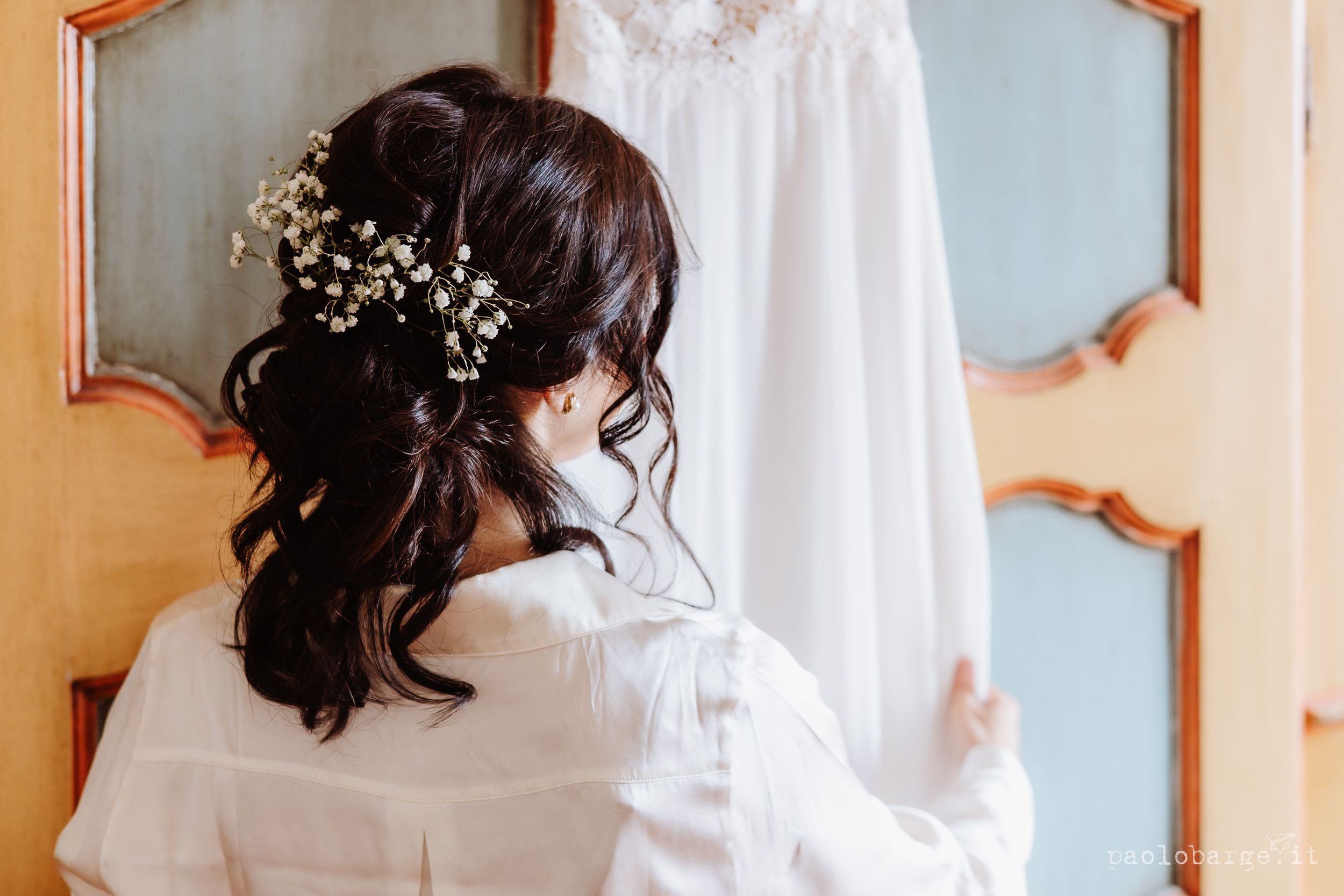 Someone adjusts details in a lace wedding dress preparation scene with floral decor and mirrors.