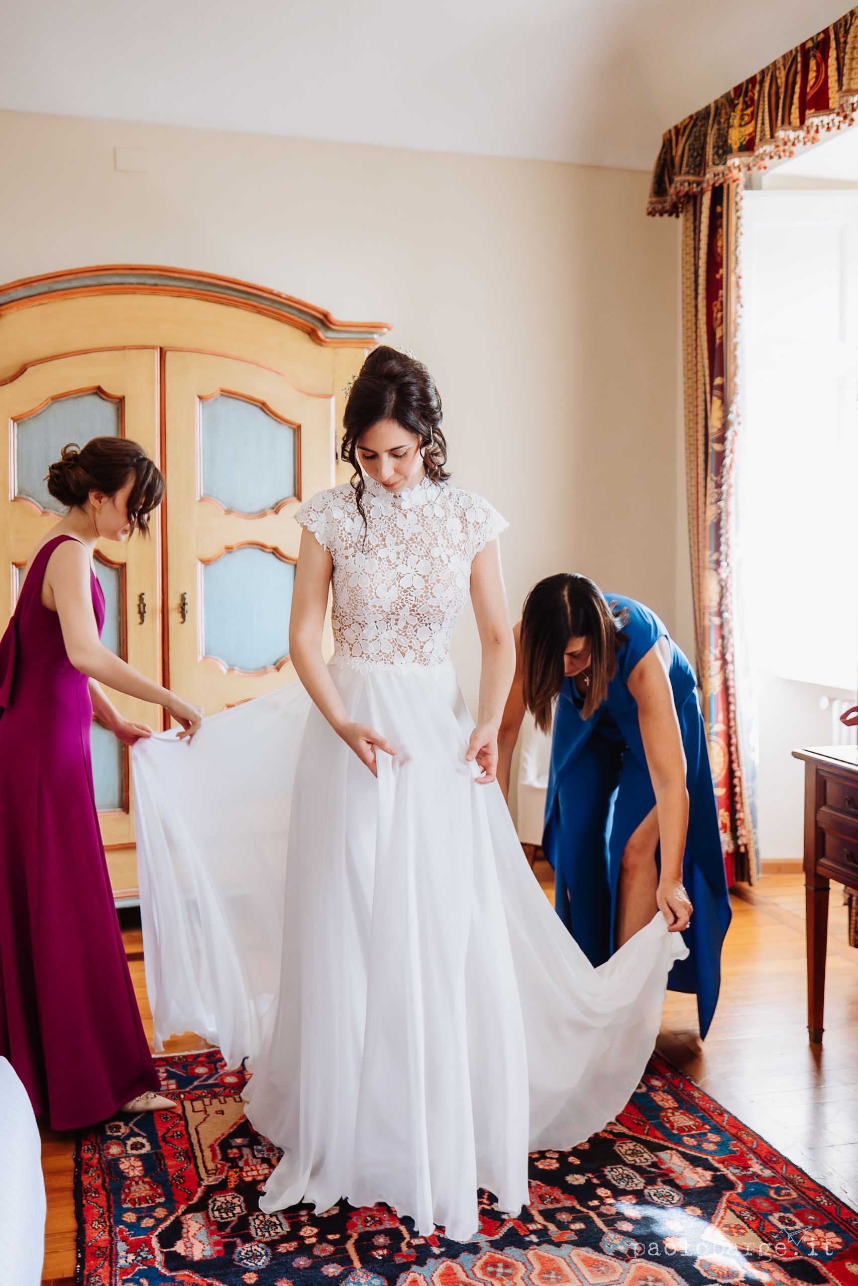 Two attendants in burgundy and blue dresses help adjust a flowing white wedding gown on an ornate rug in a bedroom.