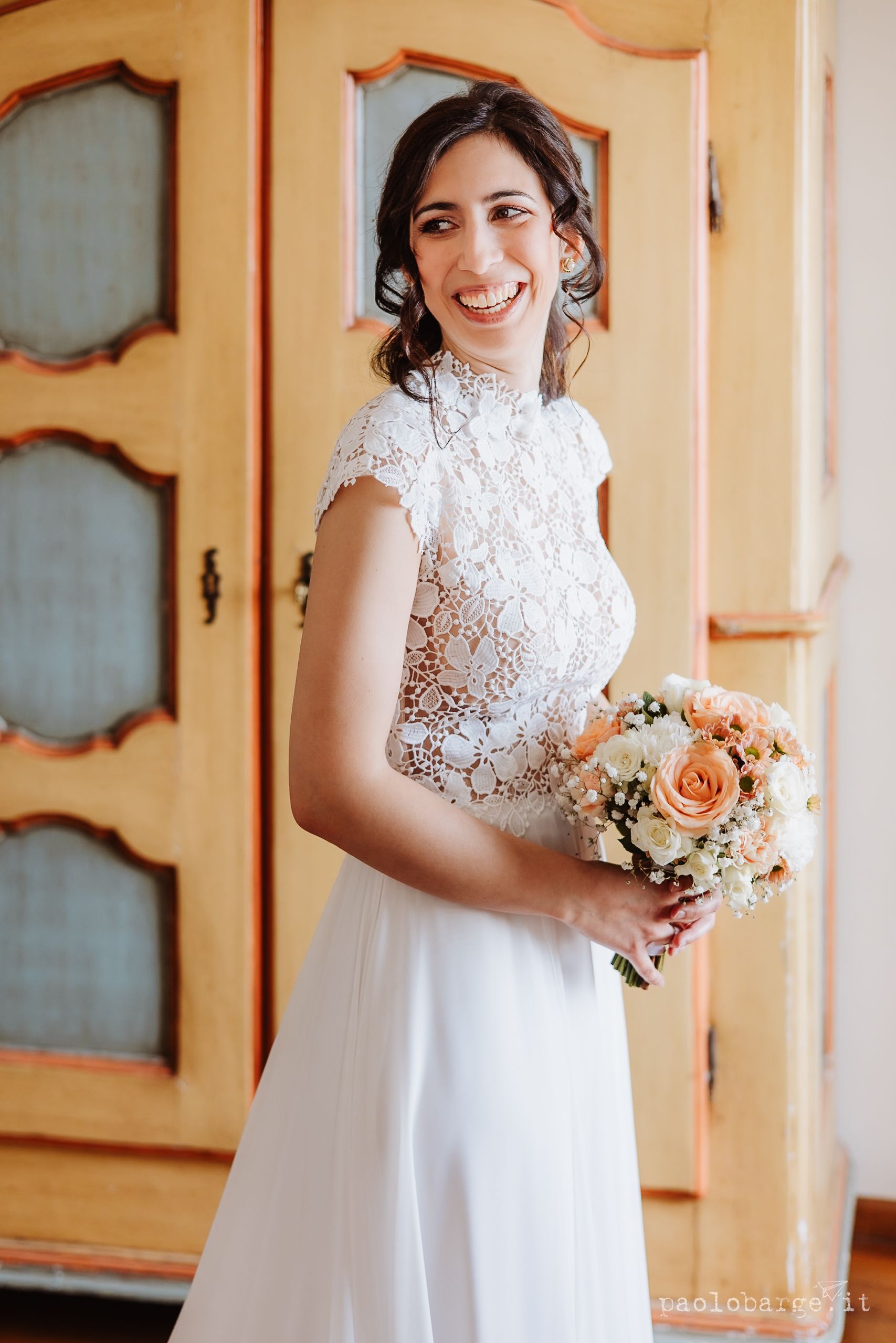 A bride wearing an elegant lace high-neck wedding dress smiles while holding a peach and white floral bouquet.
