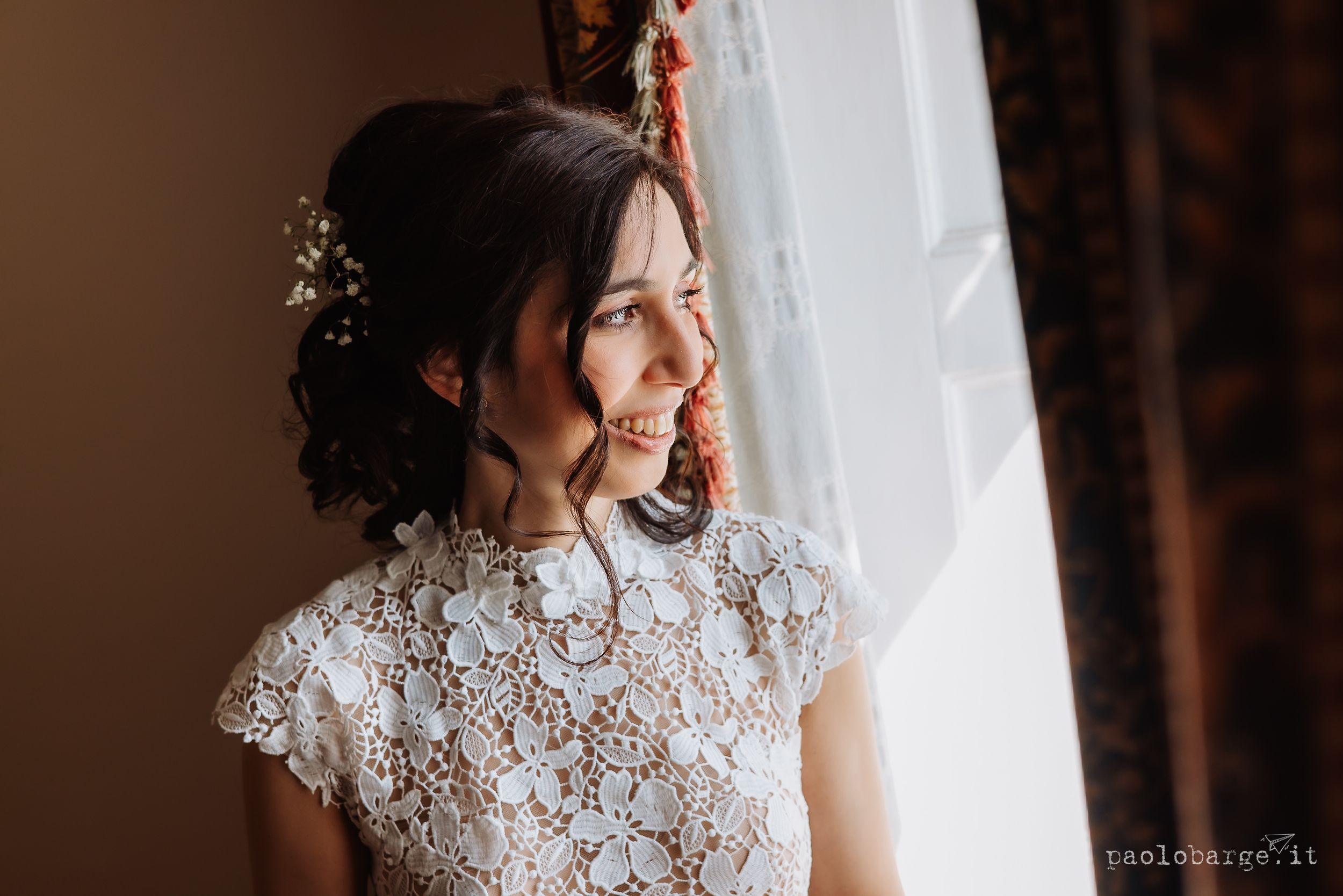 Side profile of a bride wearing an elegant white lace wedding dress with high neck detail, smiling by a window.