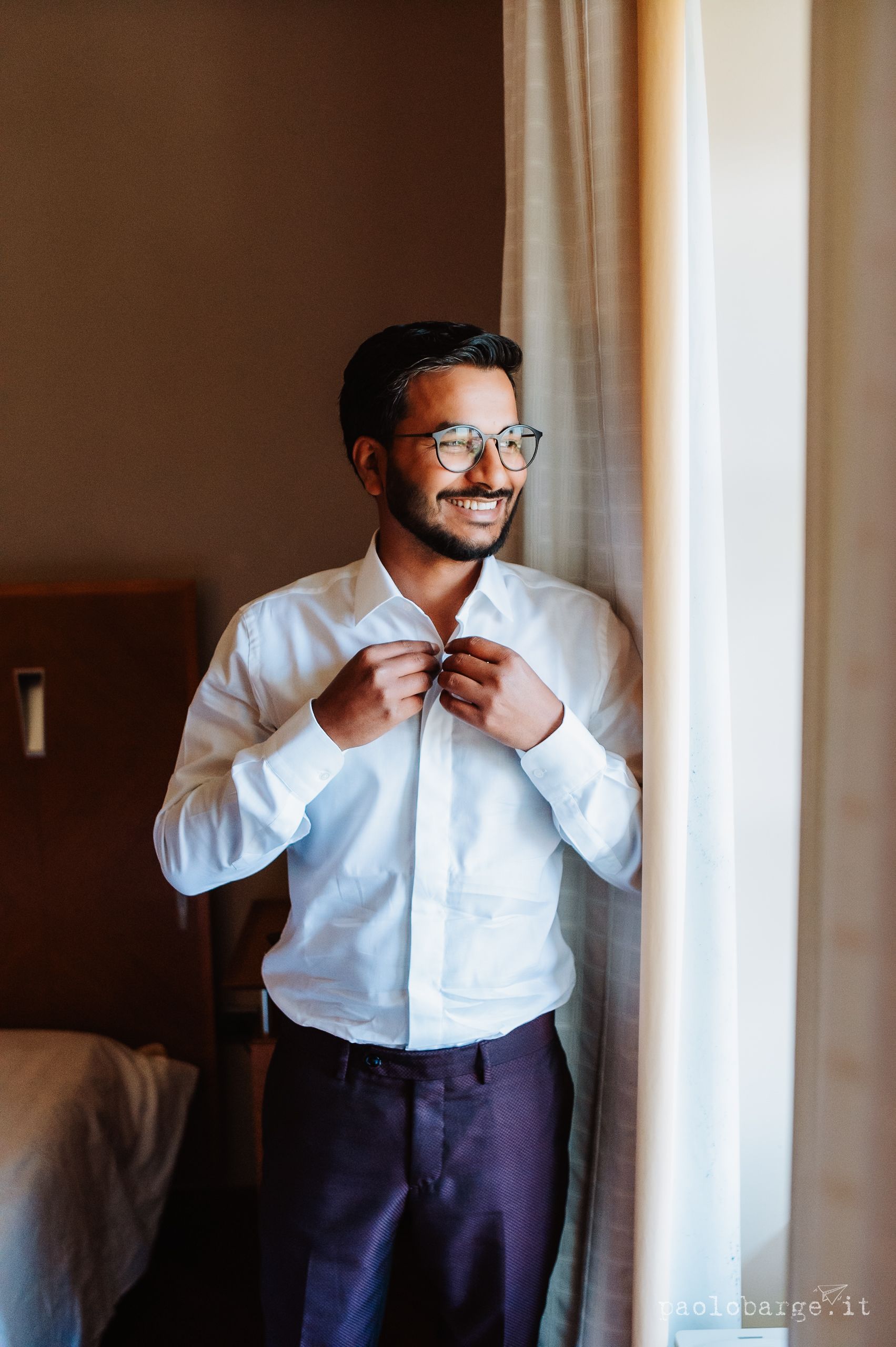 Man adjusts his shirt collar while standing next to a window in a formal white dress shirt and dark pants.