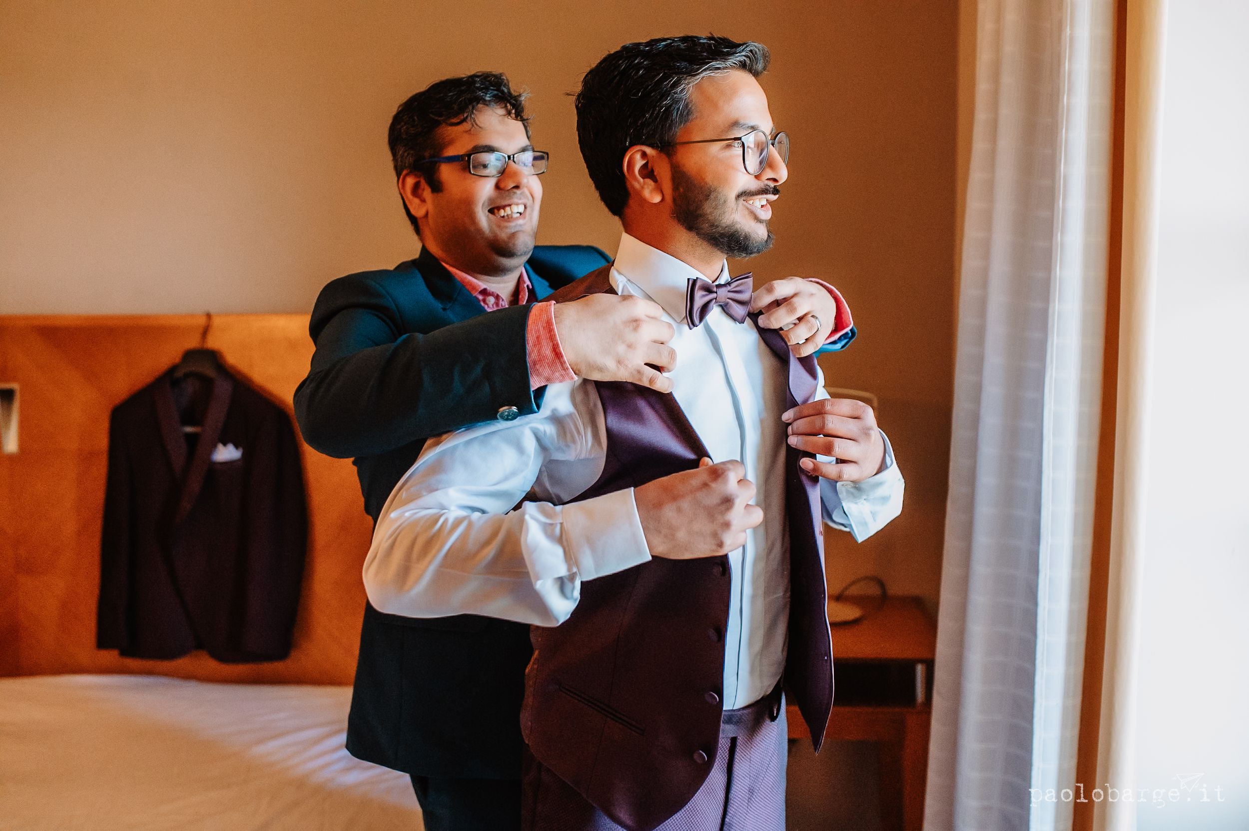 Two people in formal attire preparing before an event, with one helping the other adjust their bowtie.