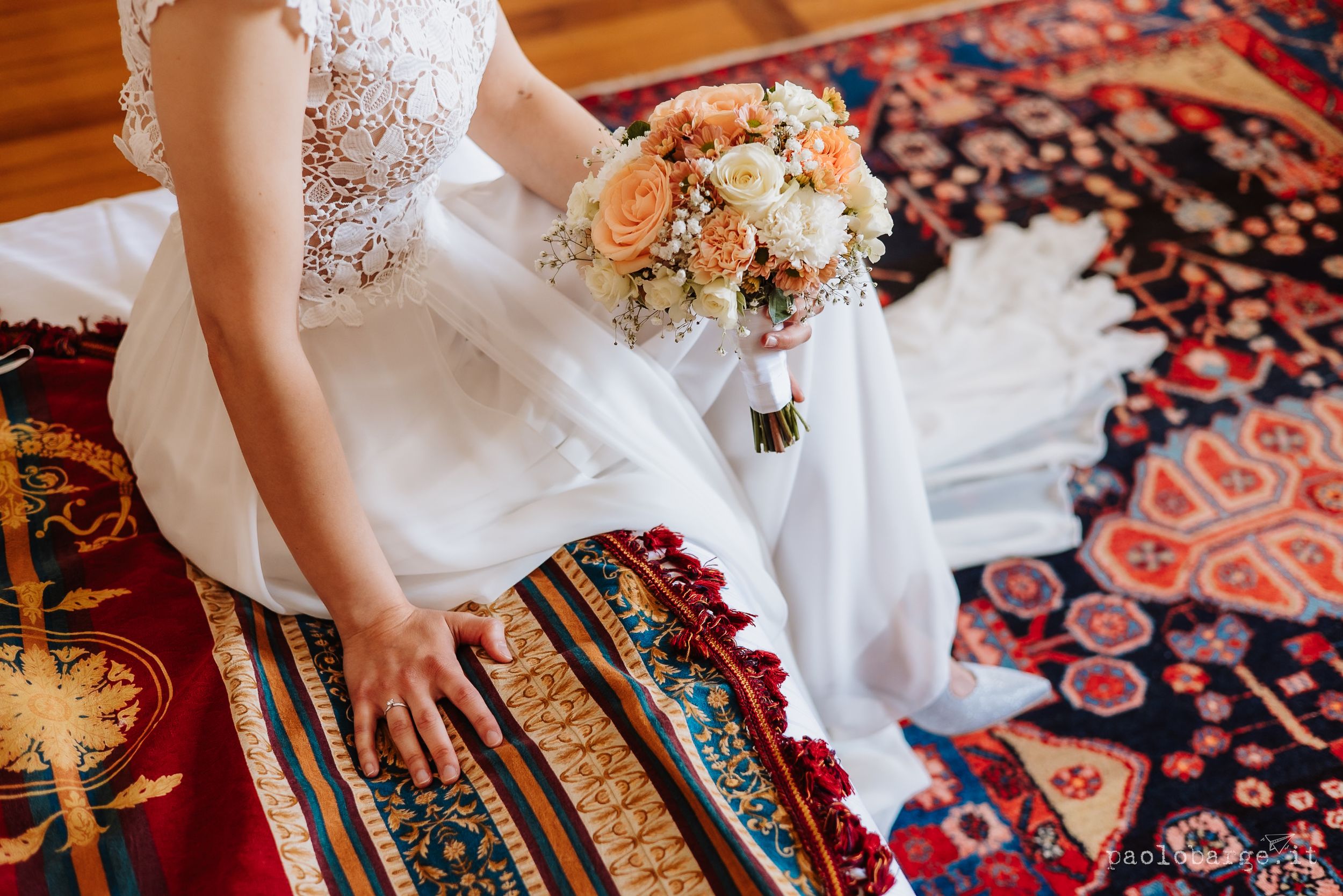 A wedding bouquet with peach and white flowers rests beside flowing white bridal gown on colorful vintage rug.