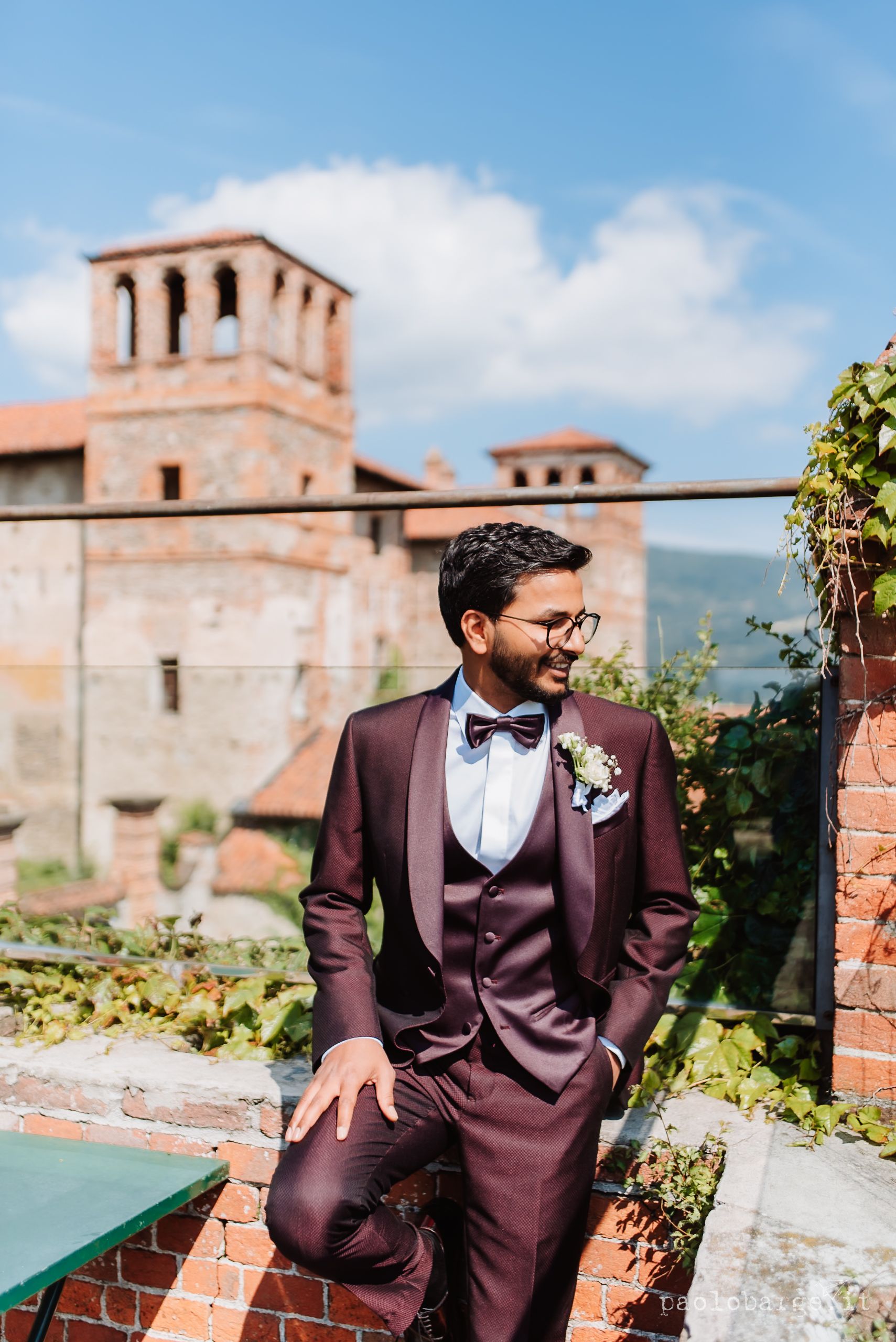 Burgundy three-piece wedding suit with bowtie photographed against Italian villa backdrop on sunny day.