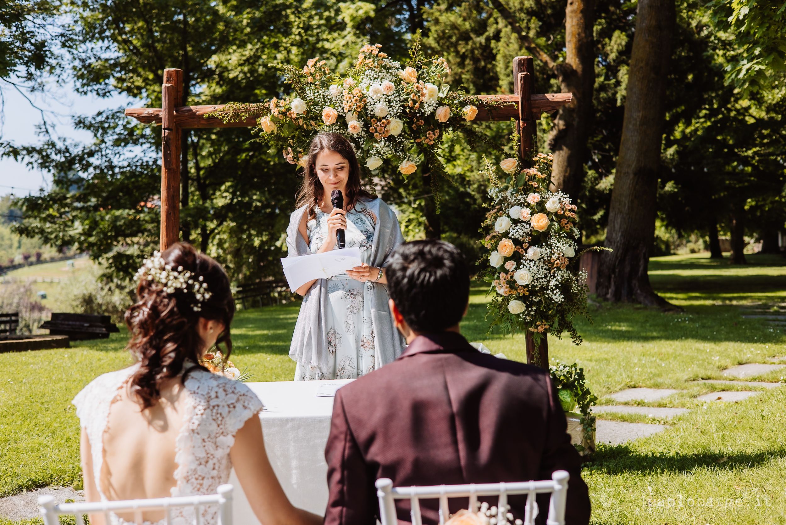 Outdoor wedding ceremony under wooden arch decorated with white and peach floral arrangements on sunny summer day.