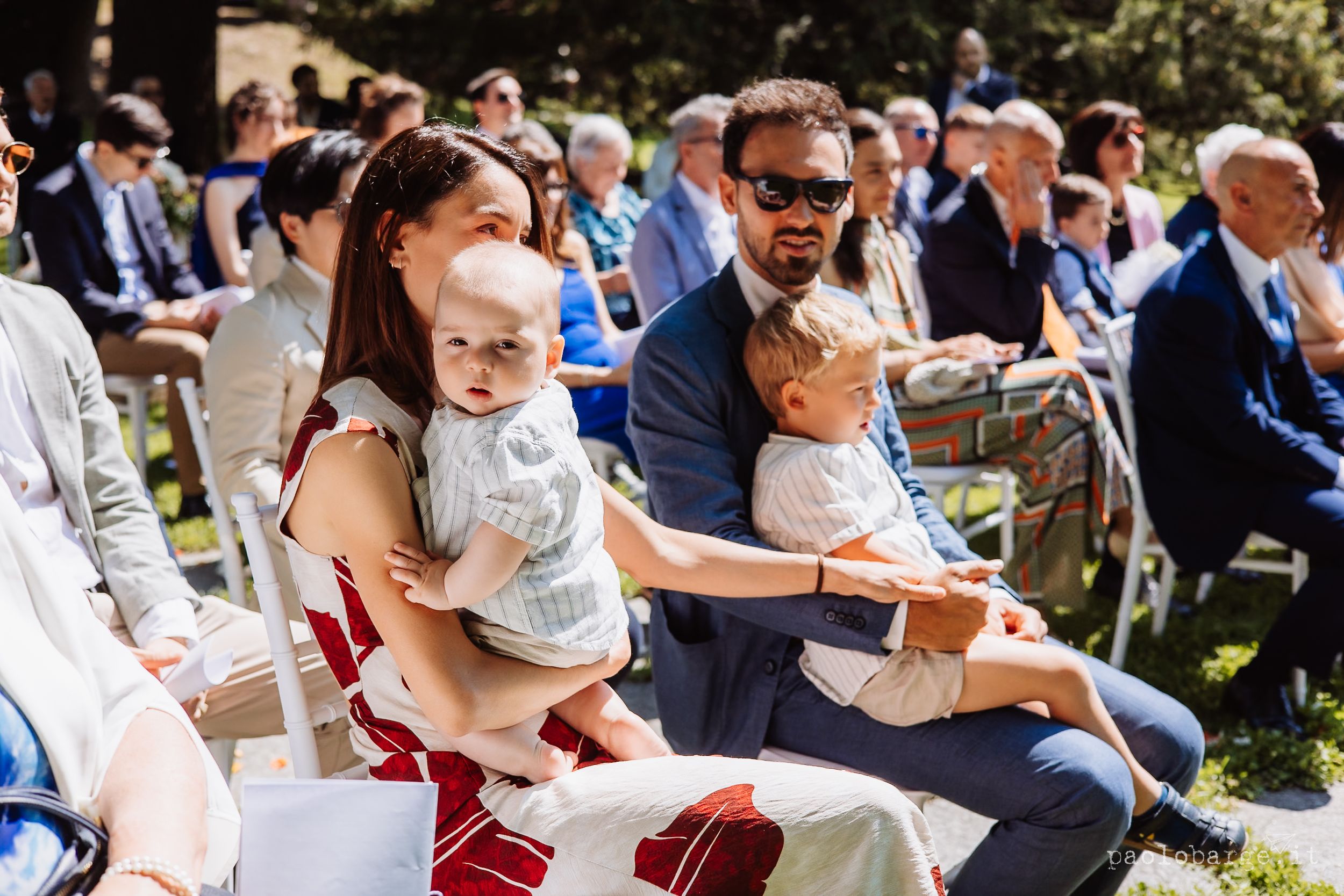 Wedding guests sit outdoors in white chairs during an elegant garden ceremony on a sunny afternoon.