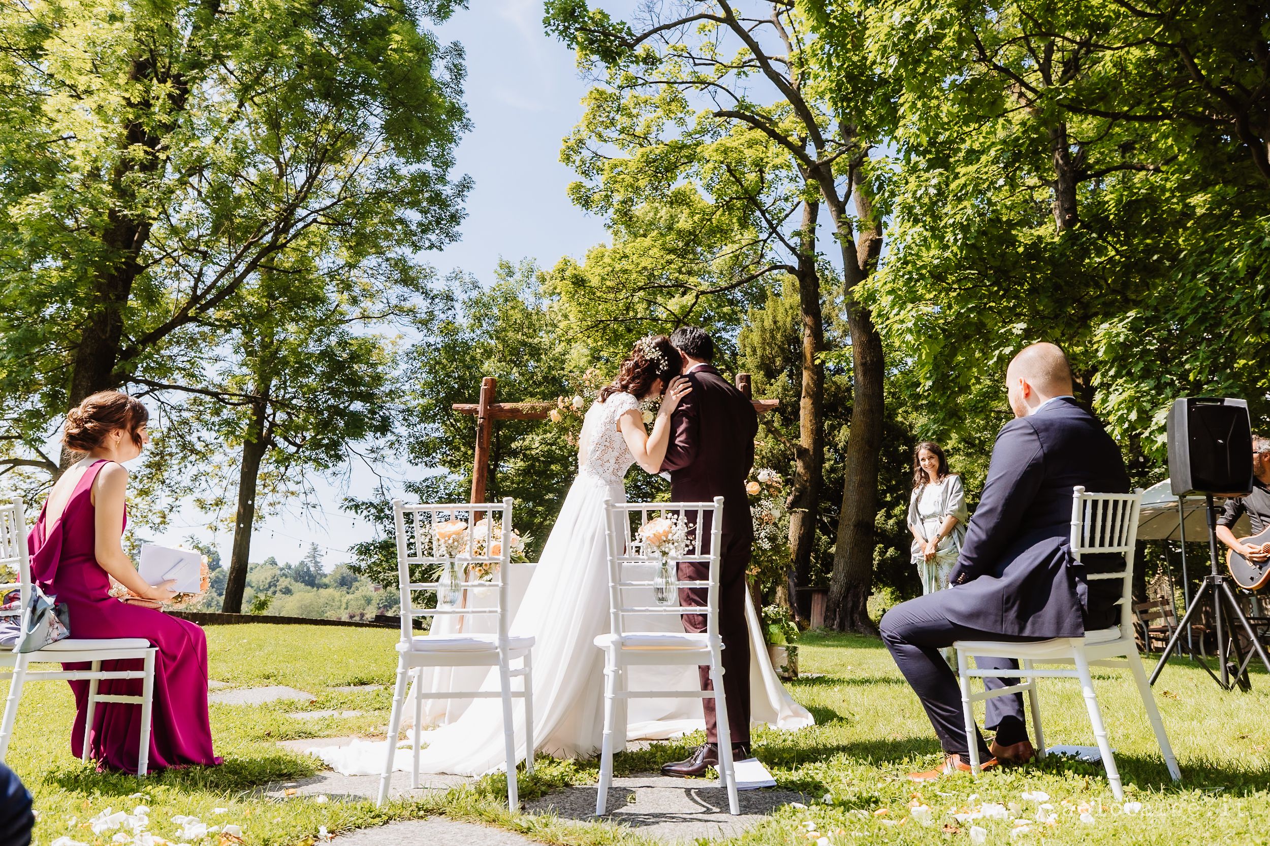 A sunny outdoor wedding ceremony with guests seated in white chairs surrounded by lush green trees and grass.