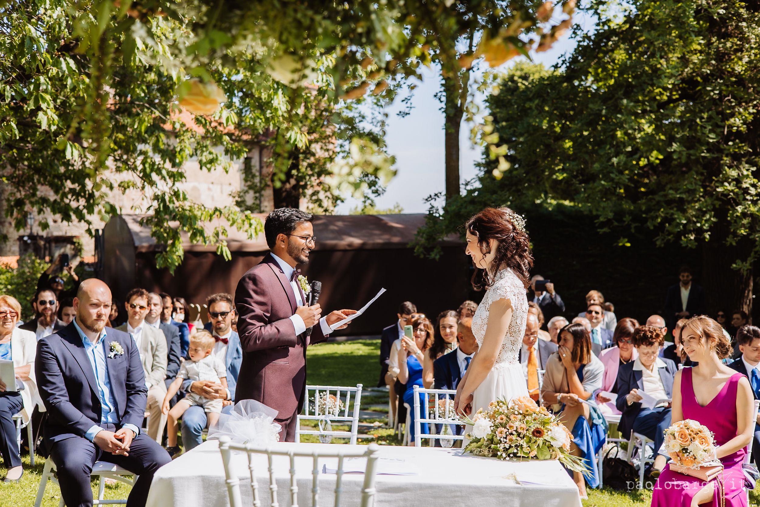 Wedding guests gather outdoors under trees for a ceremony with white chairs arranged in rows during a sunny day.