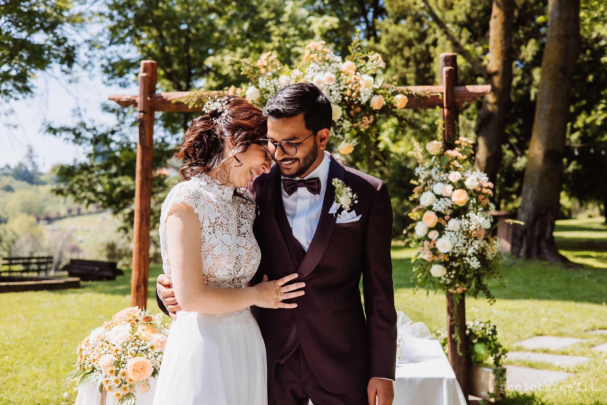 Couple embraces in a garden setting with floral archway and natural sunlight filtering through trees in the background.