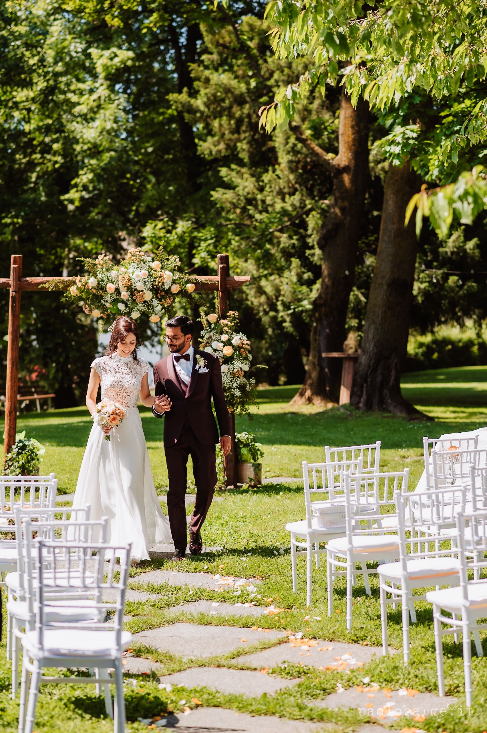 White chairs line a garden path leading to a rustic wooden arch decorated with flowers at an outdoor summer wedding ceremony.