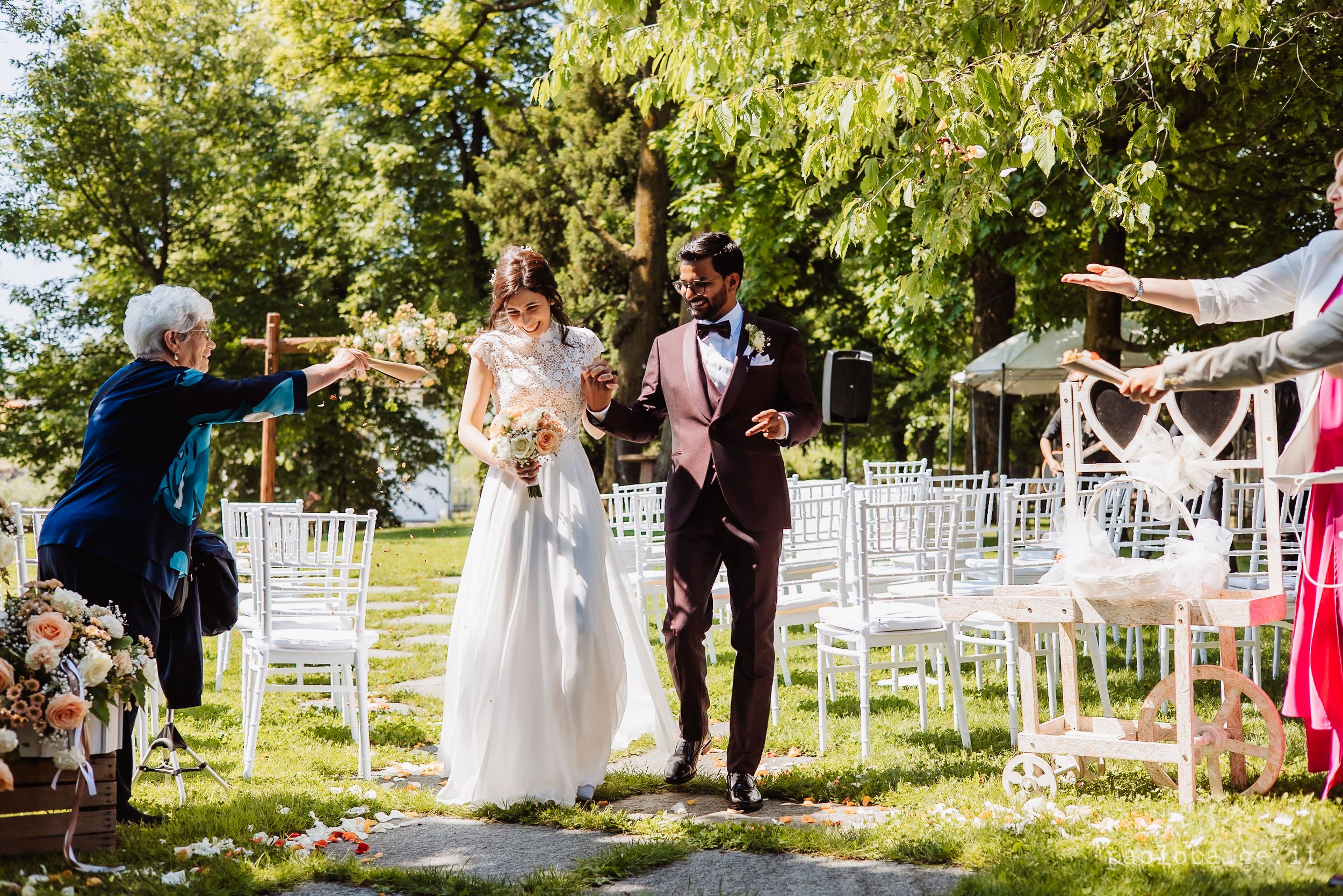 A joyful outdoor wedding ceremony with white chairs set up on a lawn beneath leafy green trees on a sunny day.