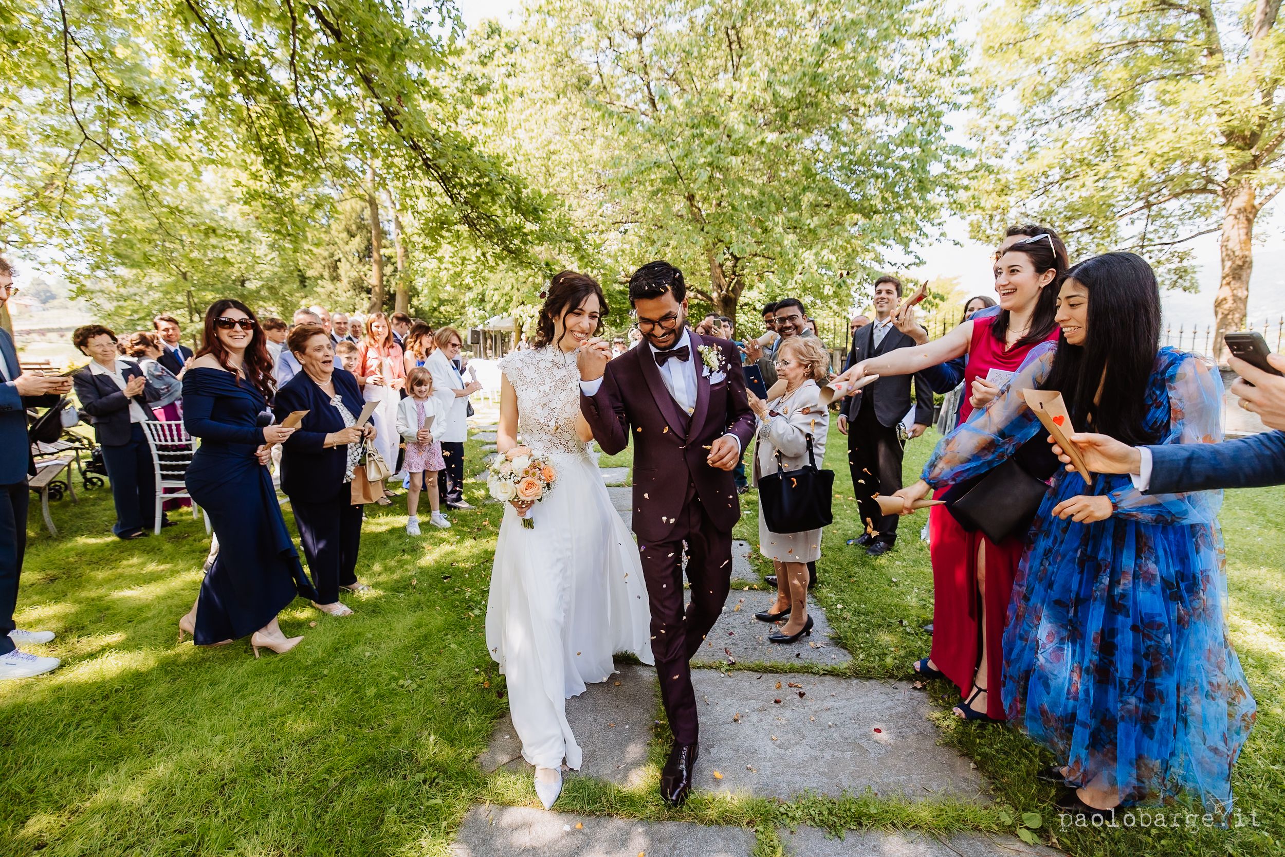 Wedding party celebrates on sunny garden lawn as newlyweds walk past smiling guests during outdoor ceremony.