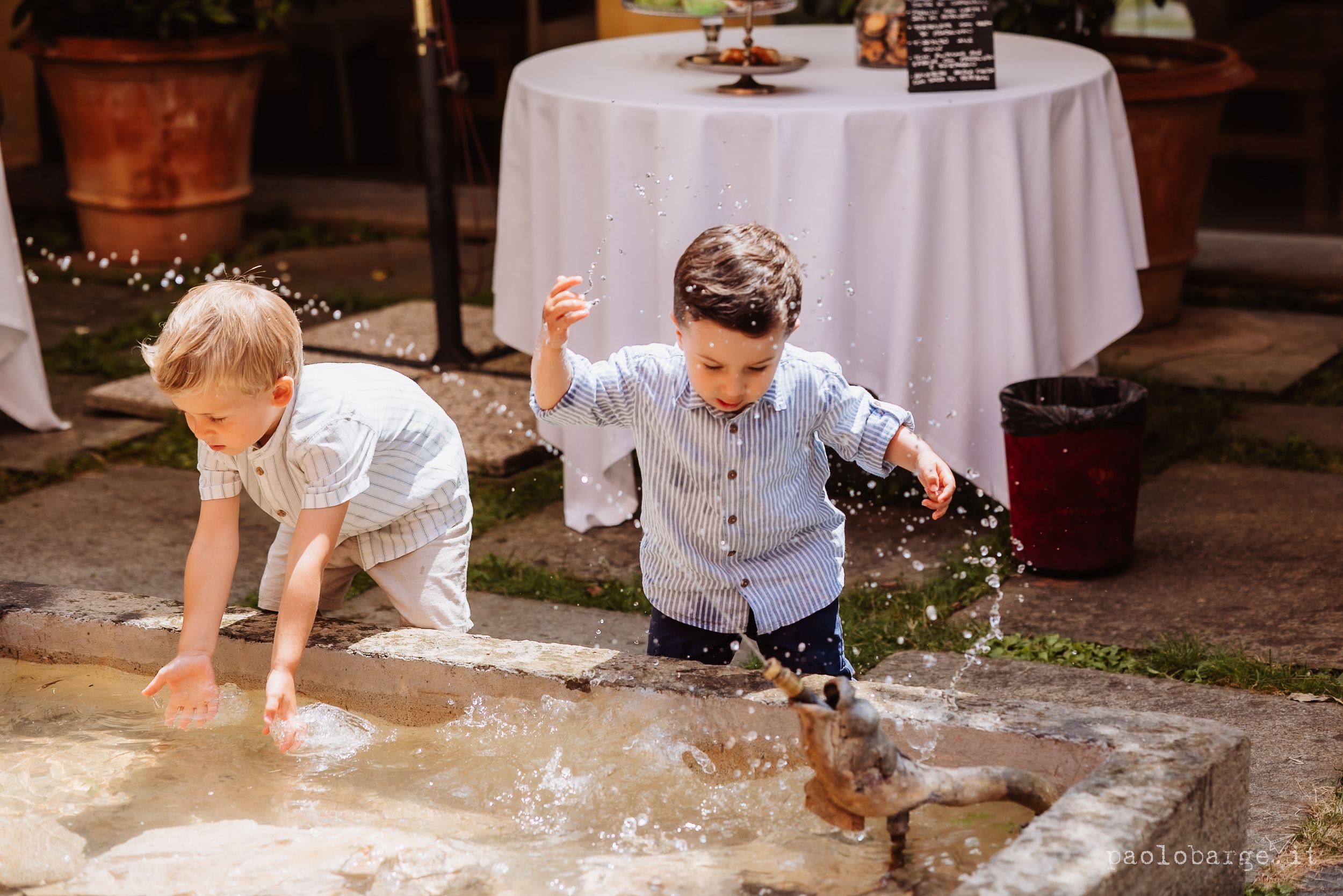 A toddler splashes and plays in a puddle of water during an outdoor event with others in the background.