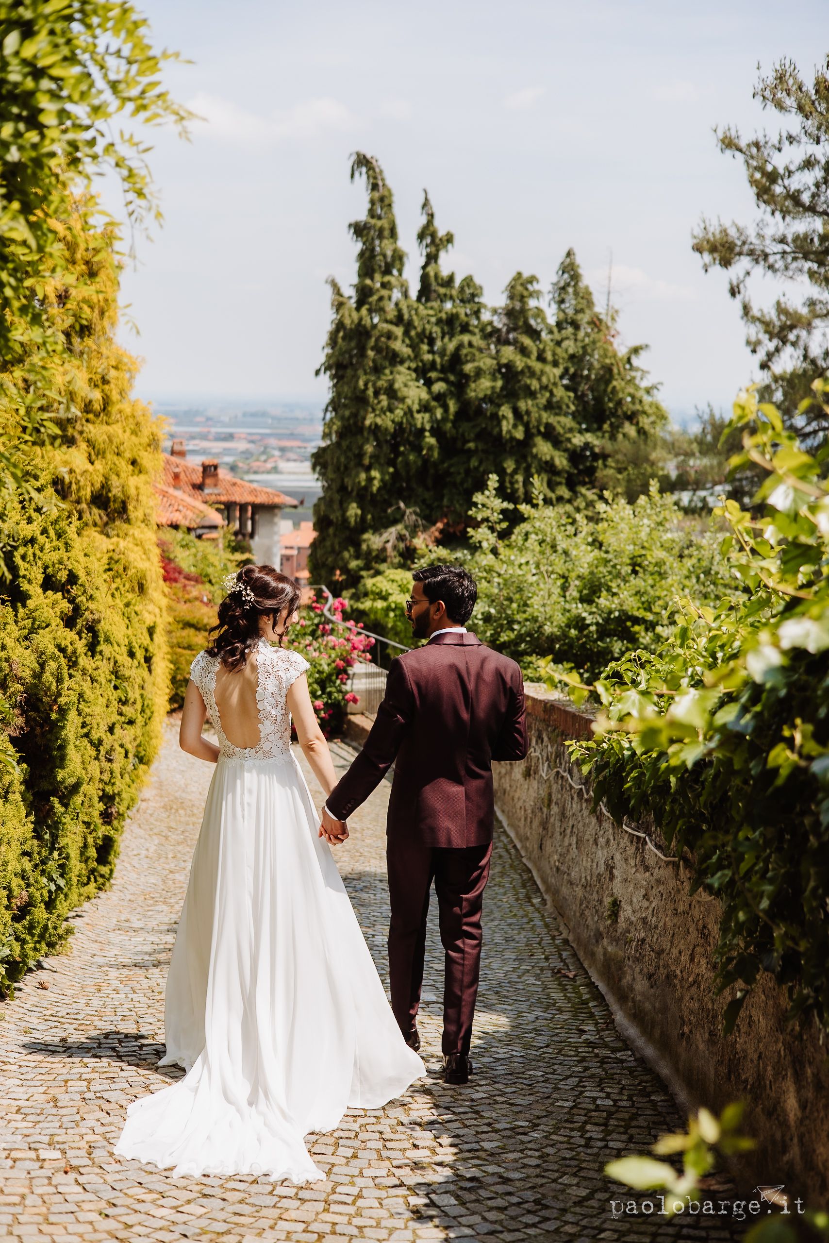 A couple walking hand in hand down a cobblestone path surrounded by cypress trees and greenery in a European town setting.