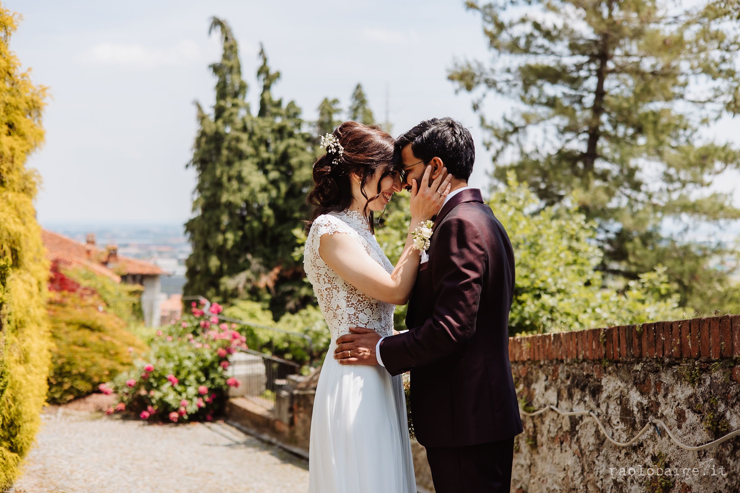 A romantic wedding couple shares an intimate moment together in a sun-drenched garden with cypress trees and stone walls.