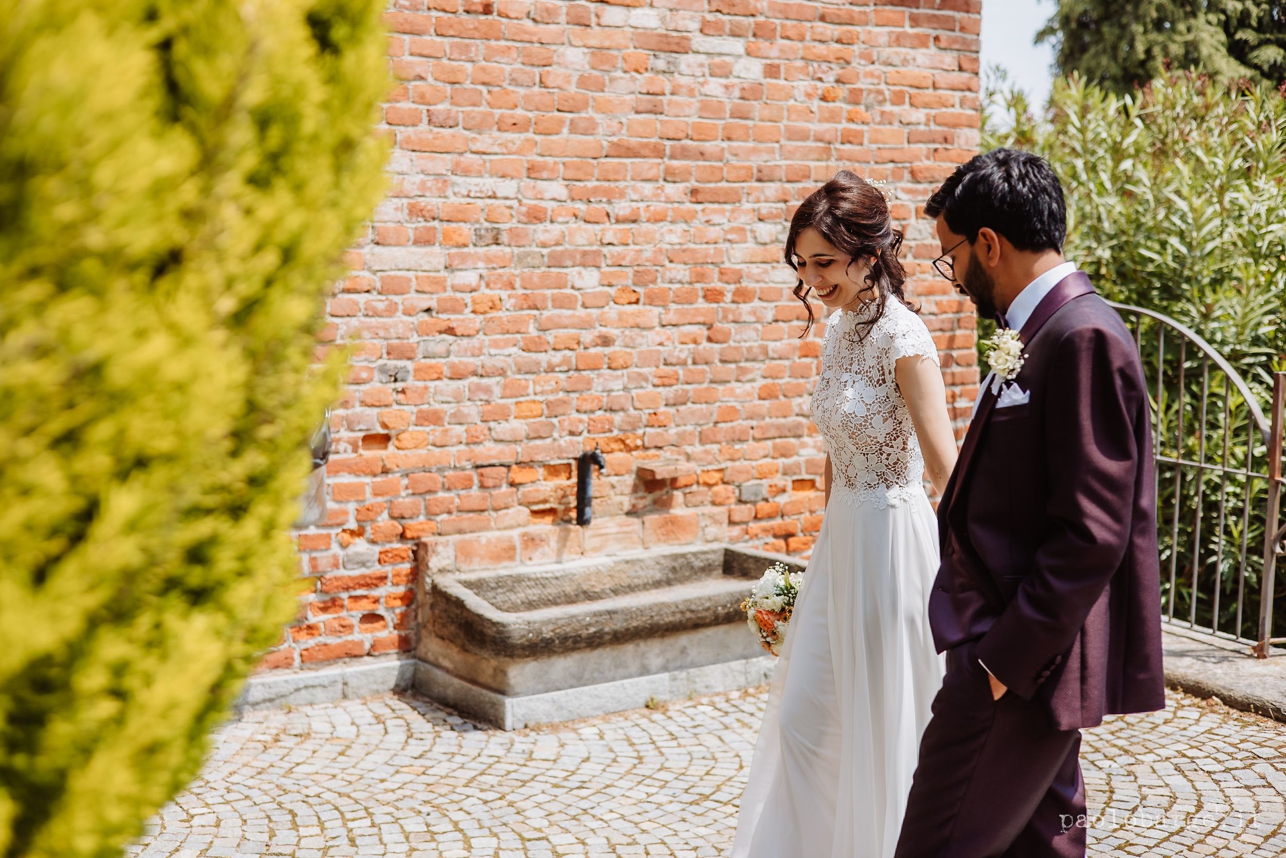 A couple in wedding attire stands near a brick wall and stone fountain on a sunny day.