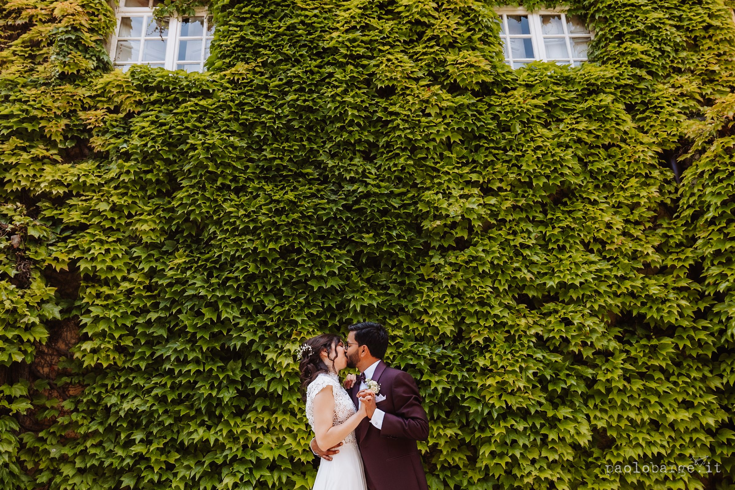 A couple shares an intimate wedding kiss in front of a lush green ivy-covered wall on their special day.