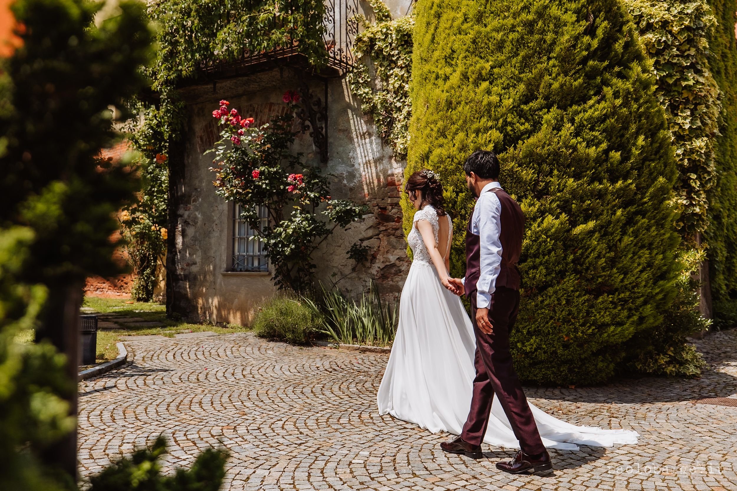 Two people walk along a scenic garden path with stone pavers surrounded by lush greenery and blooming roses.