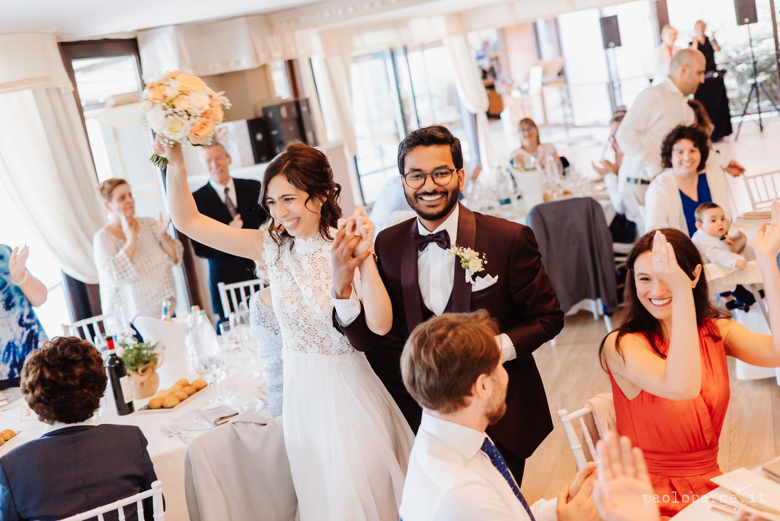 Joyful wedding guests and newlyweds celebrate during a reception in a bright indoor venue with orange floral decor.