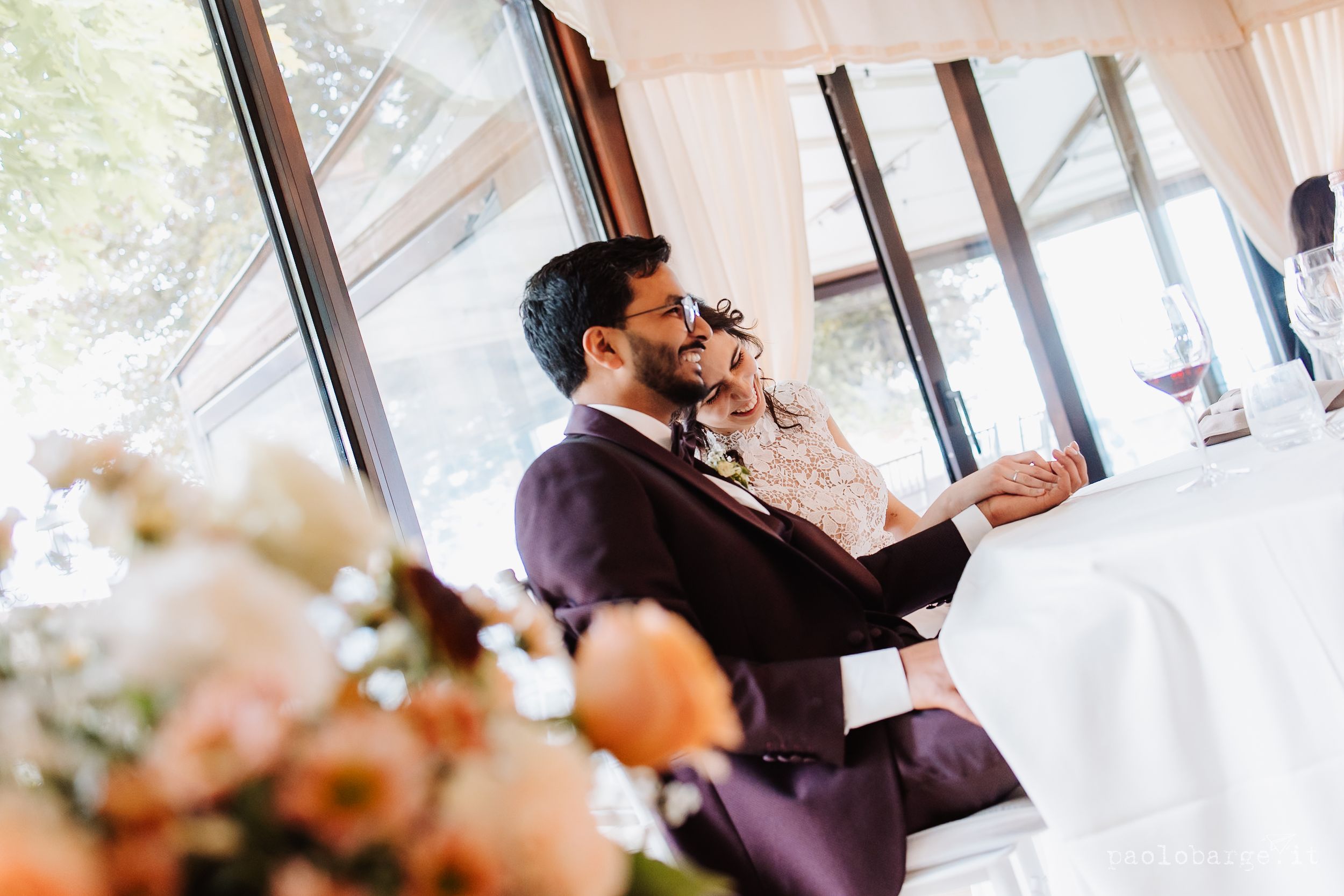 A groom in a dark suit looks out a window of a wedding venue while holding his phone to his ear.
