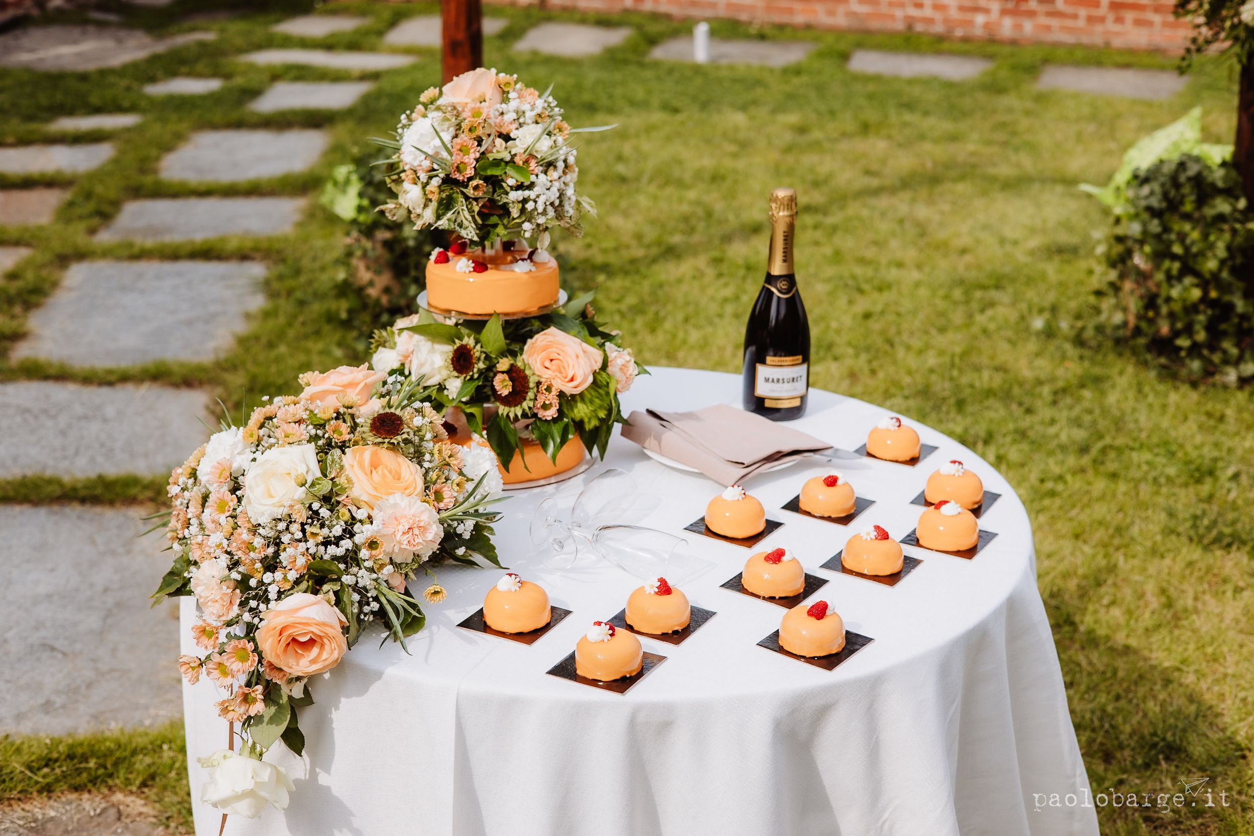 Elegant wedding table setup with heart-shaped cookies and champagne bottle on white tablecloth in garden setting.