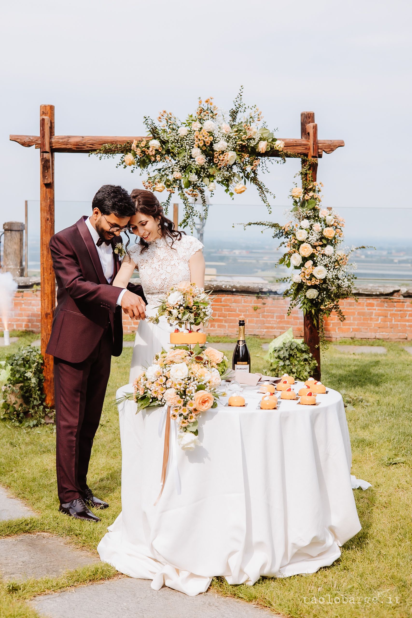 Outdoor wedding ceremony setup with wooden arches adorned with peach and white floral arrangements and white tablecloths.