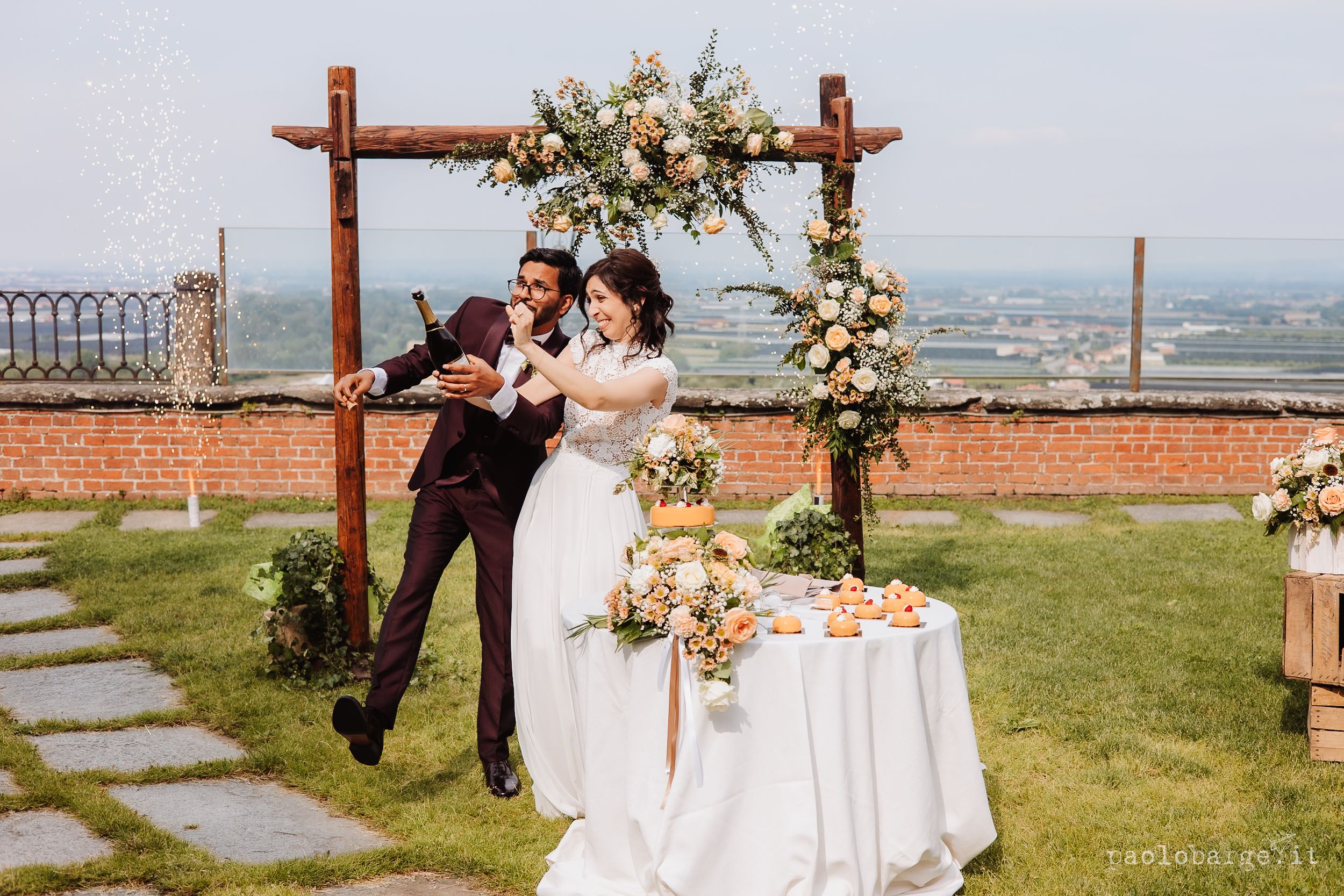 Rustic wooden arch decorated with cream flowers sits atop hilltop overlooking city during outdoor ceremony.
