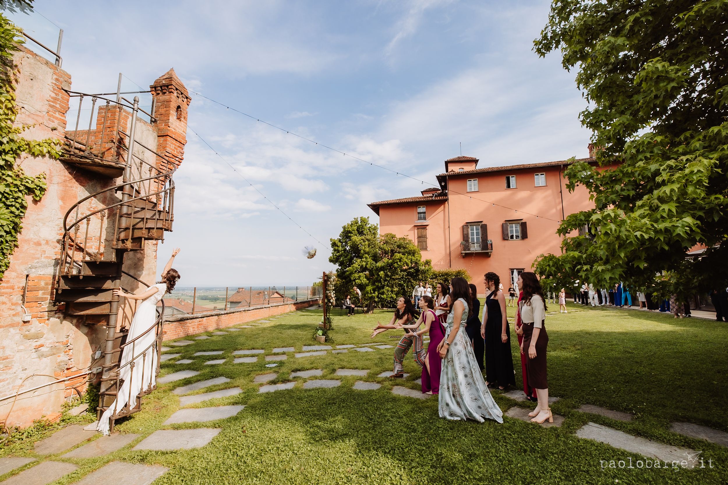 Small group of wedding guests walk along a stone path leading to a rustic brick building in the countryside on a sunny day.