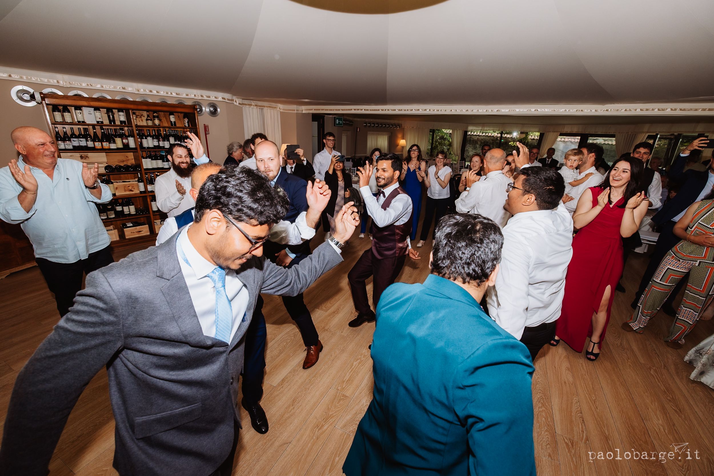 Wedding guests dance and celebrate on a wooden dance floor at an indoor reception venue during a lively party.