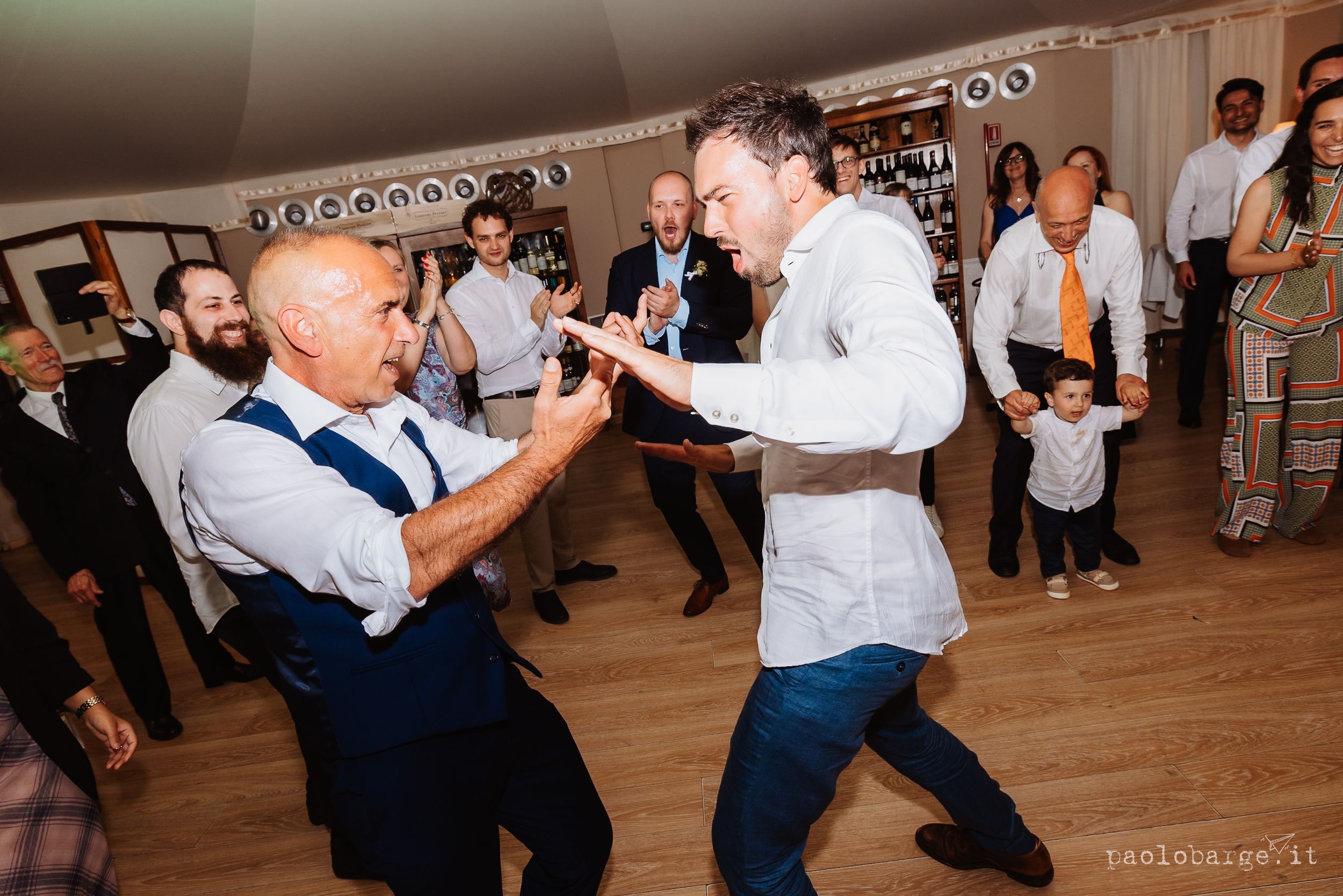 Two people enthusiastically dance together at a wedding reception on a wooden dance floor while guests look on.