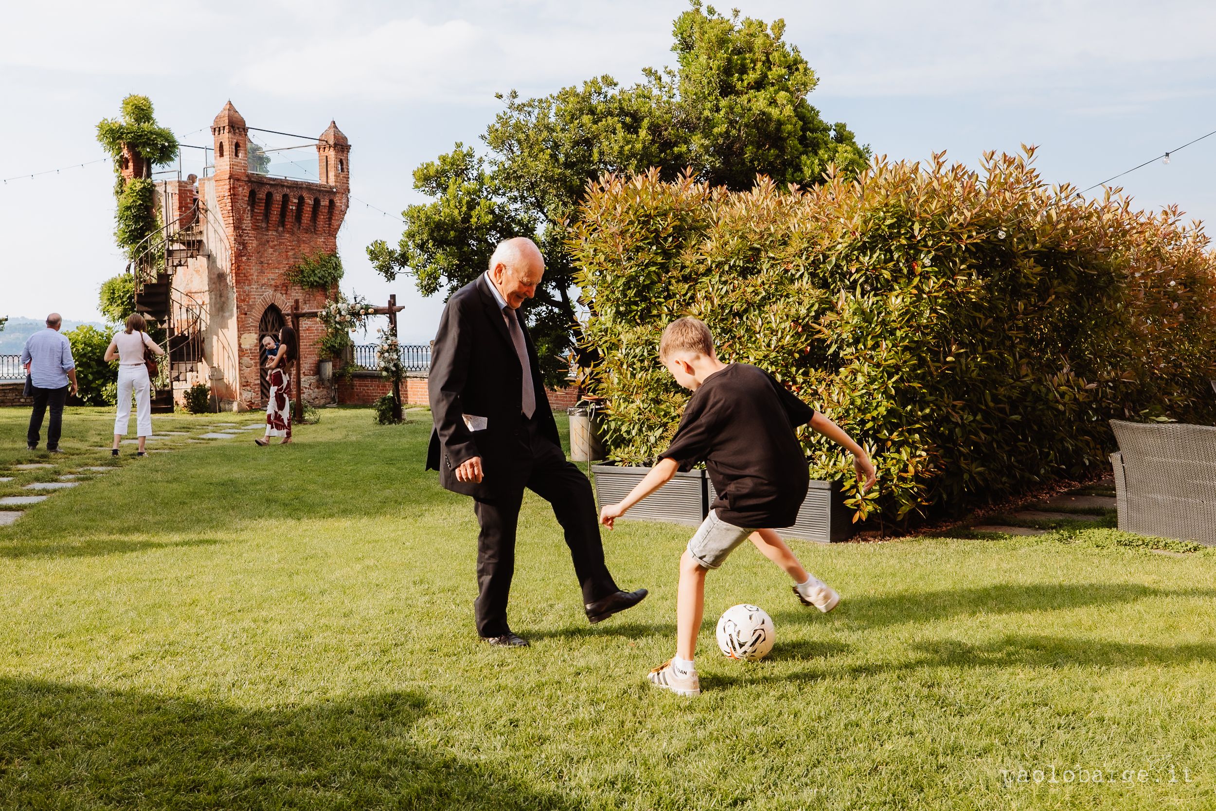 People playing soccer on a lawn with a historic brick tower and landscaping in the background.