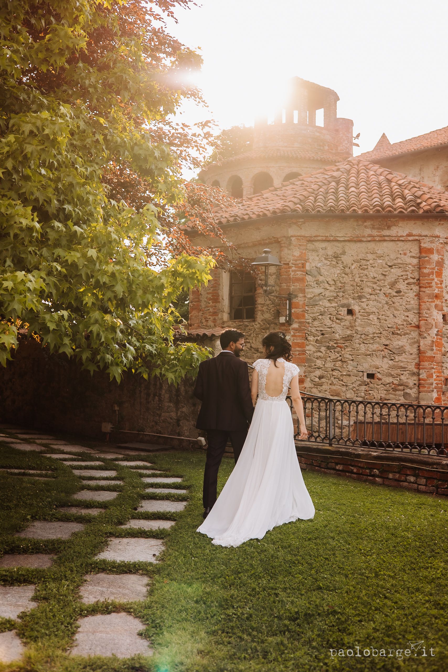 A couple walks along a stone path towards a rustic Italian villa with warm sunlight filtering through autumn trees.
